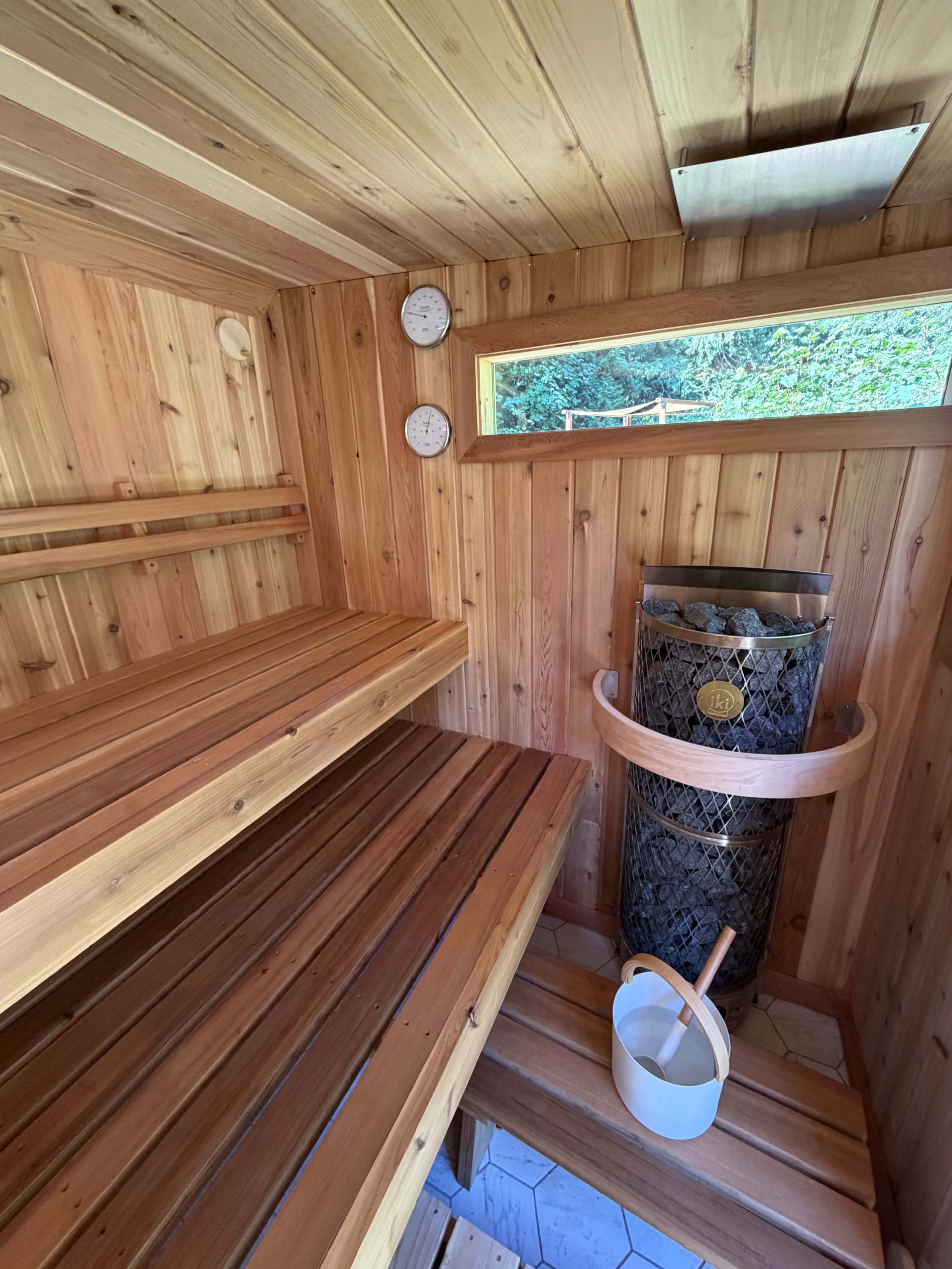 The image shows the interior of a wooden sauna, featuring slatted wooden benches, a stone-filled stove, and two thermometers or hygrometers on the wall.