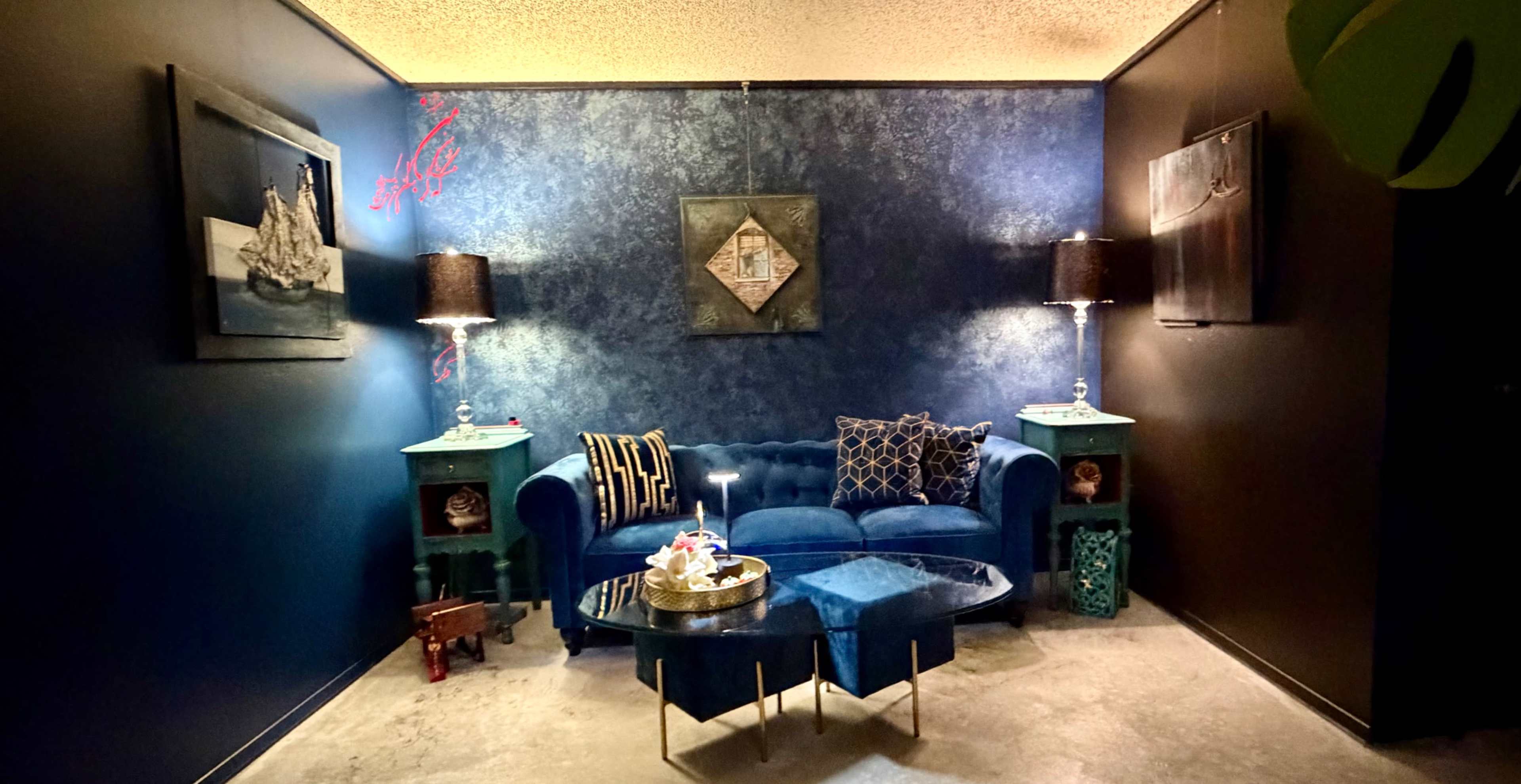 The image shows a stylish interior with a blue velvet sofa, two matching side tables, and decorative lamps, framed artwork on the walls, and a coffee table in the center.