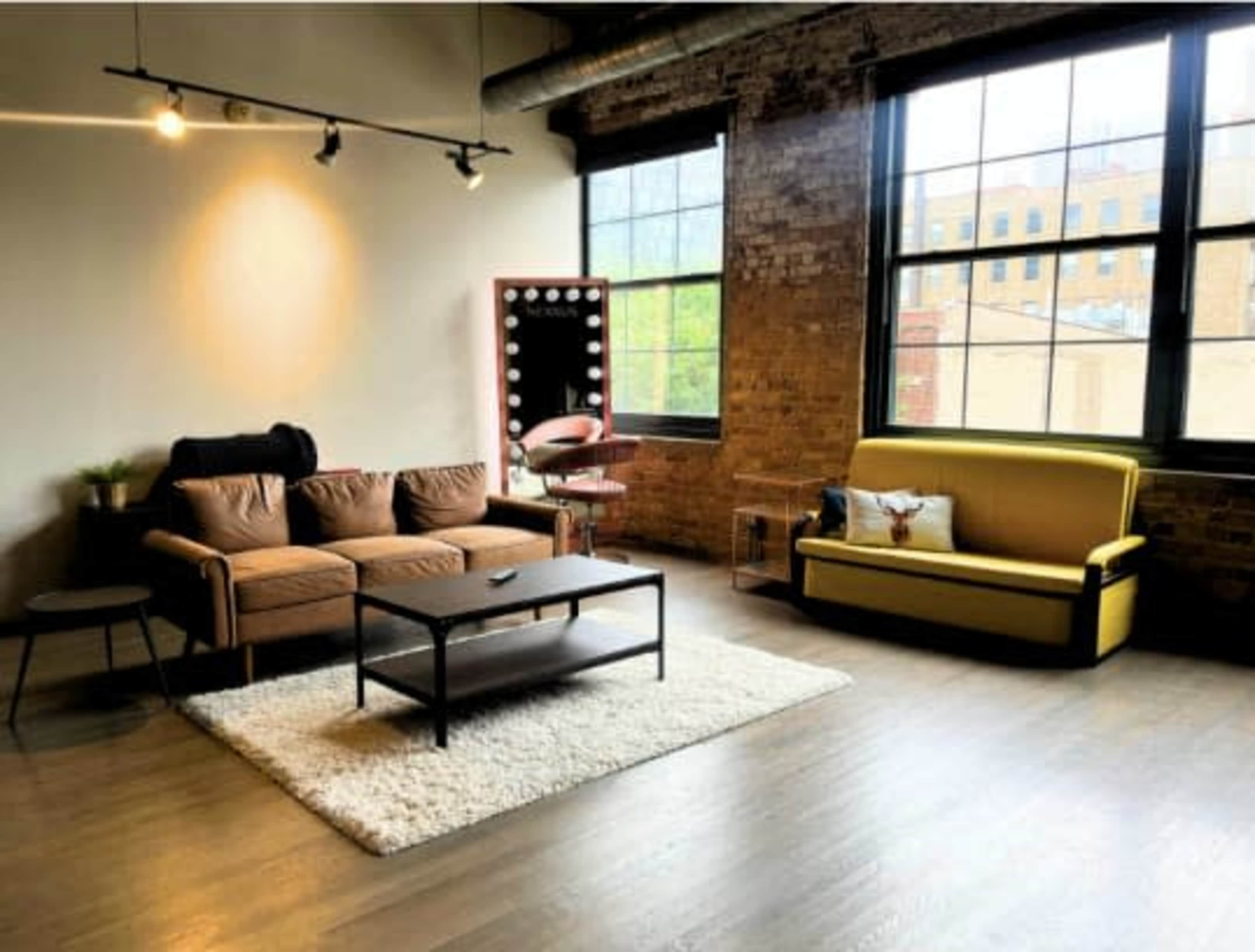 The image shows a spacious, industrial-style living area with a brown couch, a yellow sofa, a black coffee table, and large windows providing natural light.