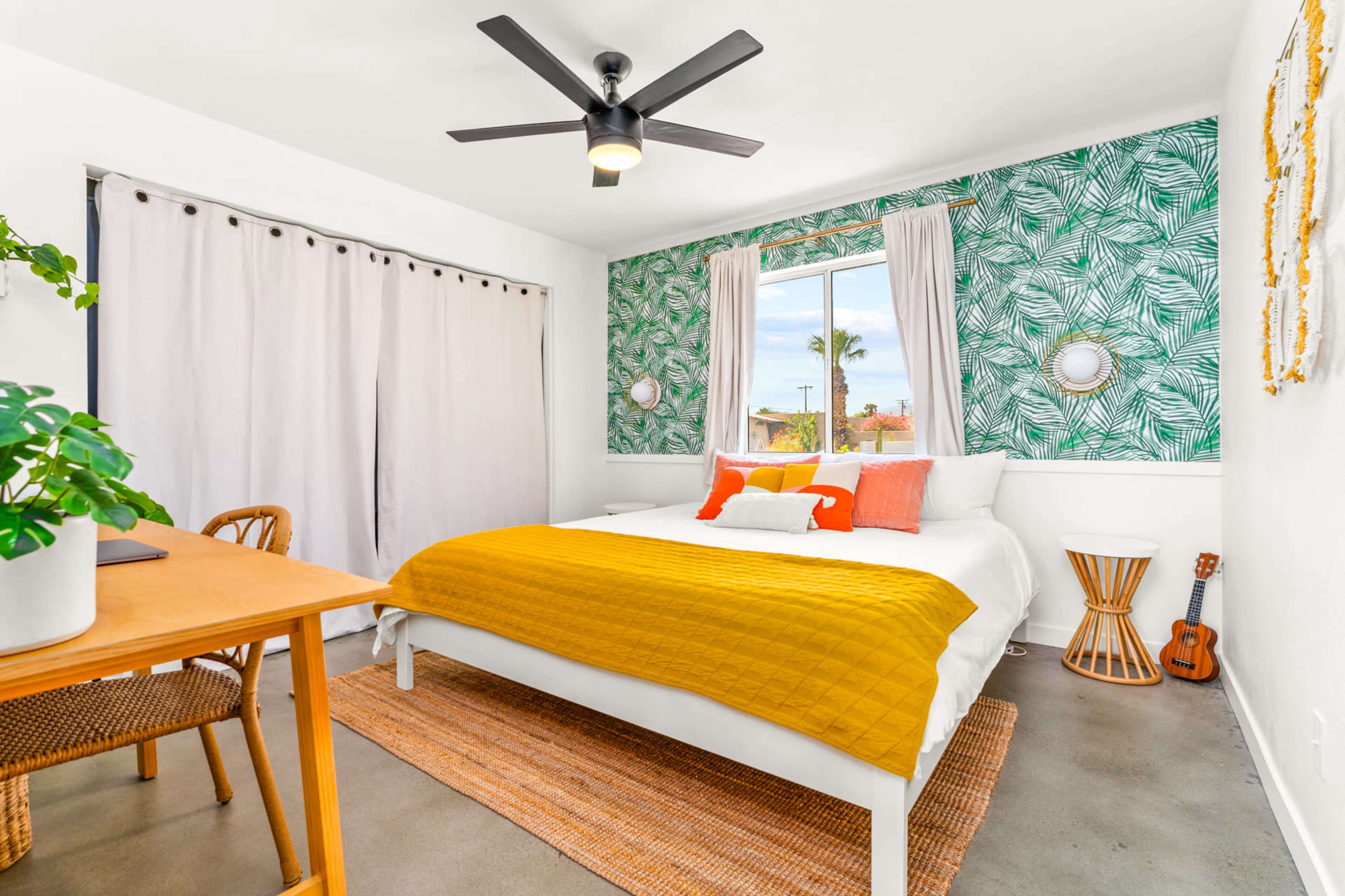 The image shows a well-decorated bedroom featuring a bed with a yellow quilt, a round table with seating, and green leaf-patterned wallpaper.
