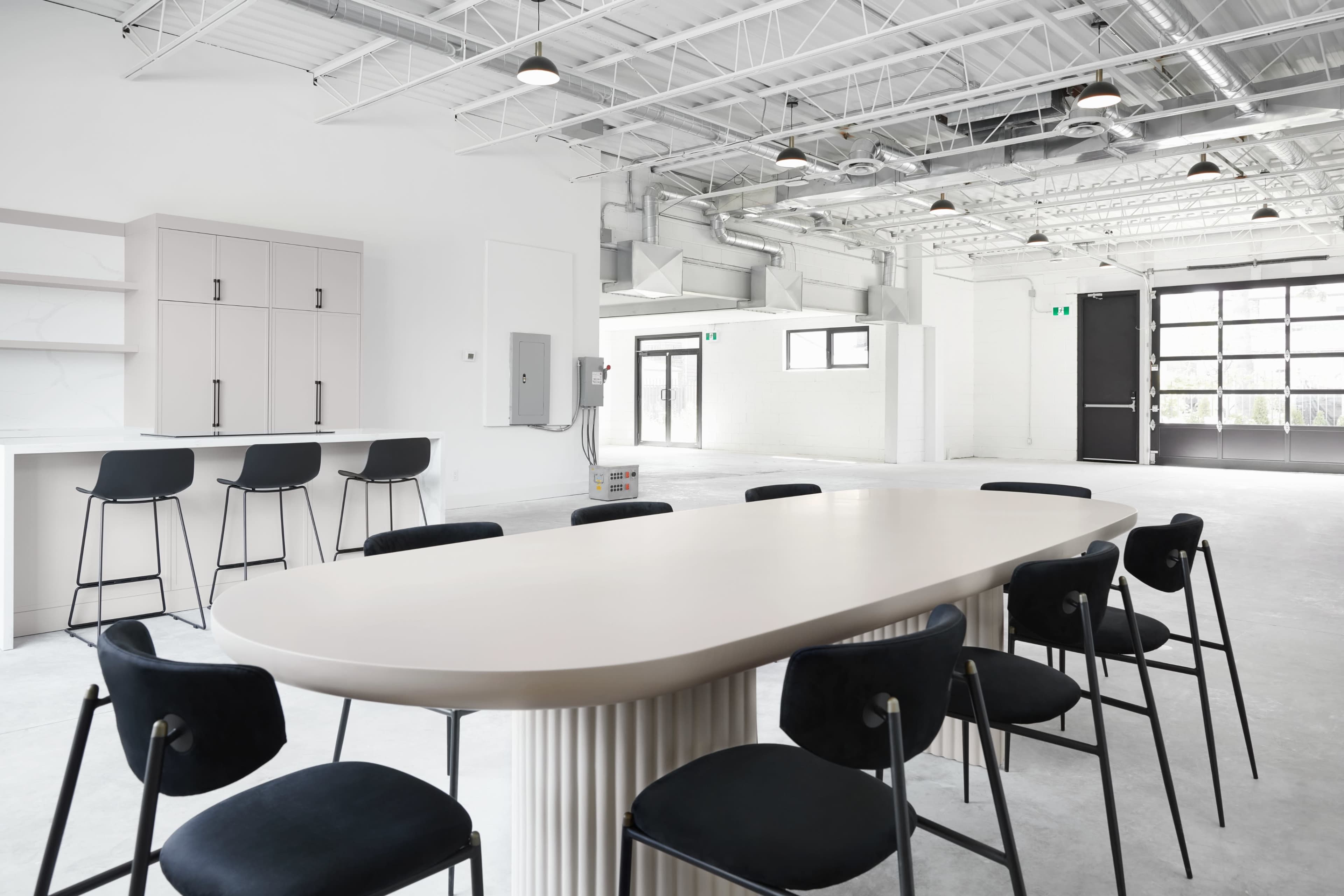 A modern, minimalist meeting space with a long oval table surrounded by black chairs, complemented by cabinetry and large windows.