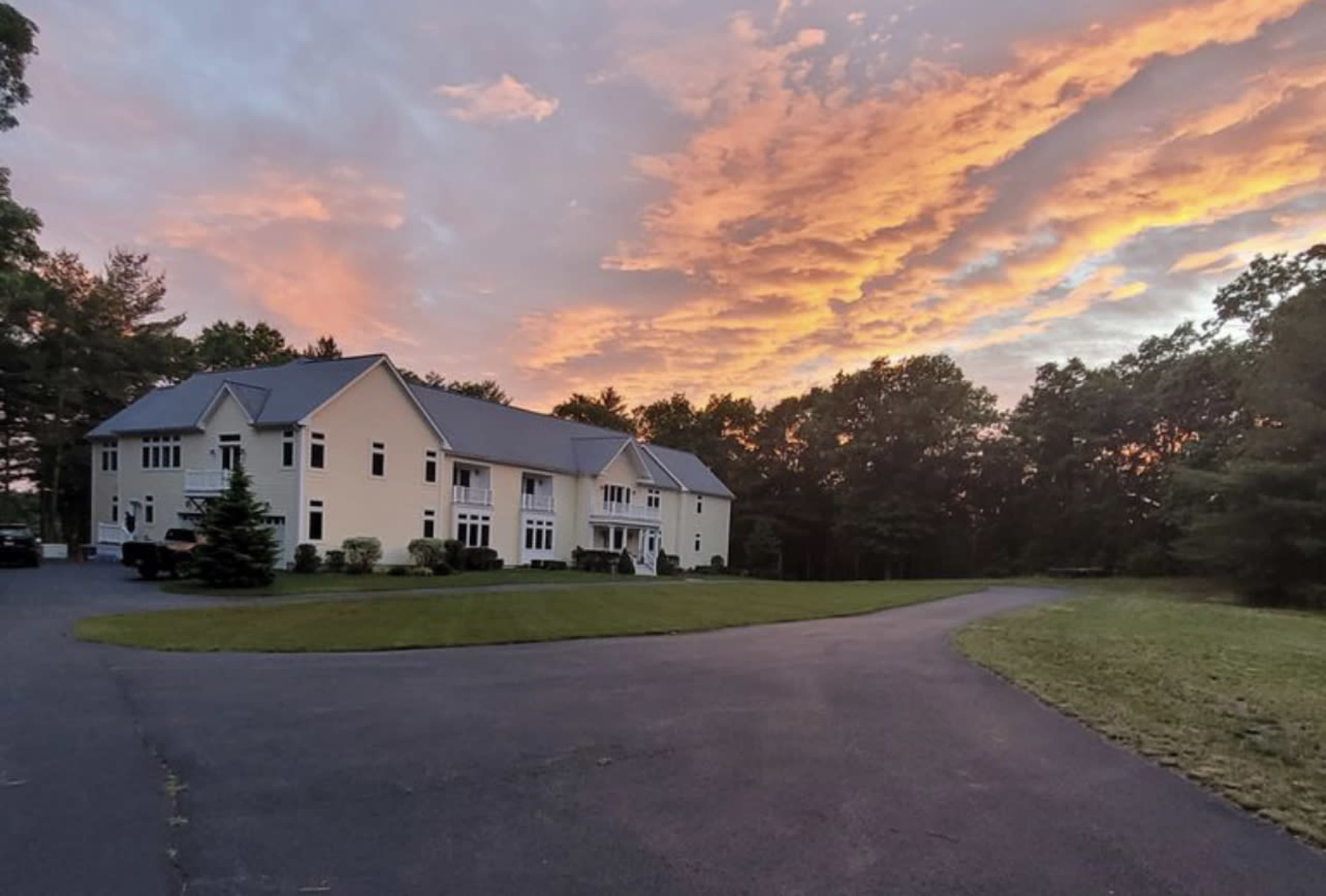 A large, two-story house with a manicured lawn is situated beside a winding driveway under a colorful sunset sky.