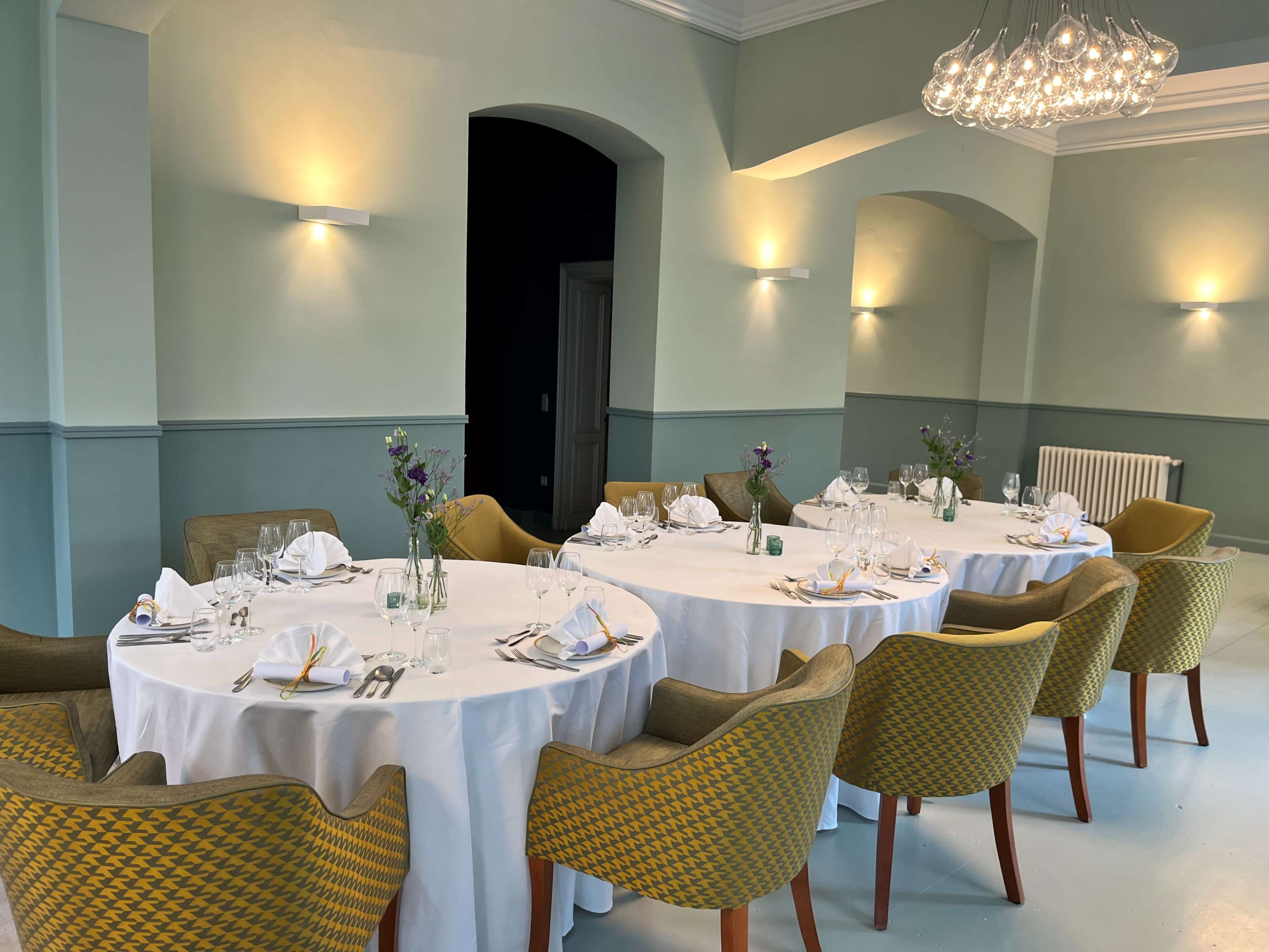A dining area with a large round table set for a meal, surrounded by green and yellow chairs, and decorated with small flower arrangements.
