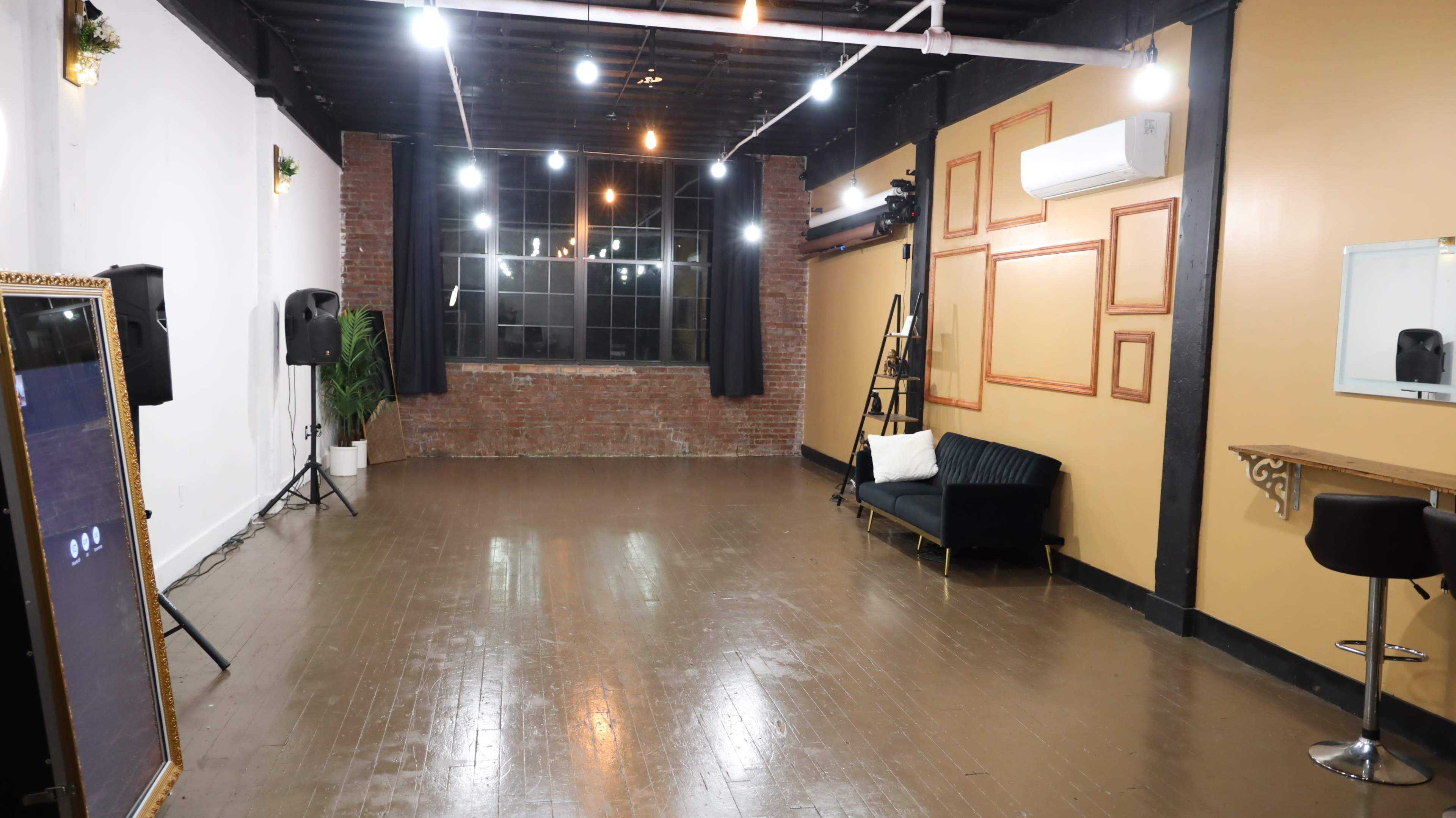 An empty indoor space with wooden flooring, exposed brick walls, a large window, and minimal furniture, including a couch and a bar-height table.
