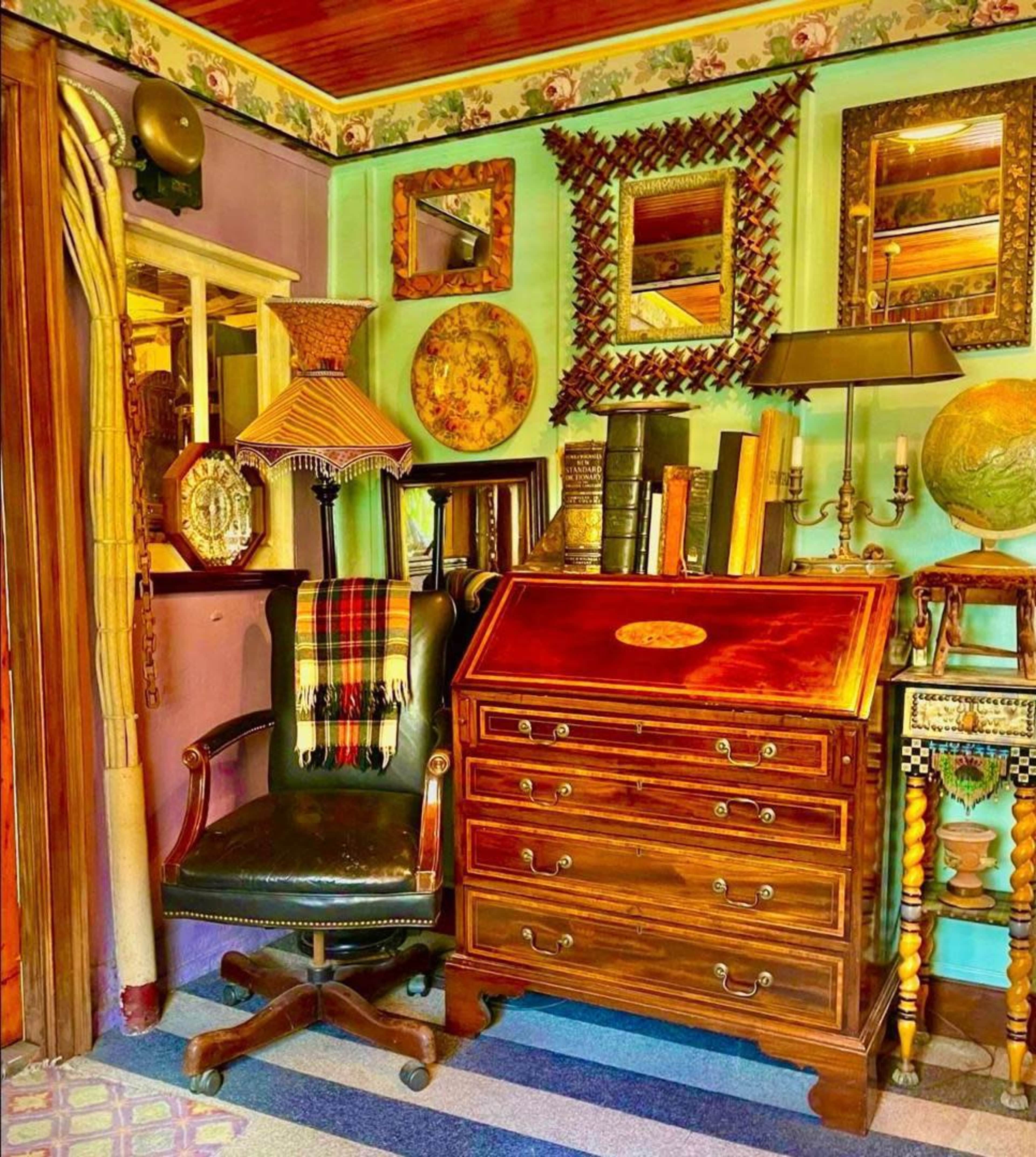 The image depicts a cozy corner of a room featuring a vintage desk, a leather chair, shelves lined with books, and various decorative mirrors and artifacts.