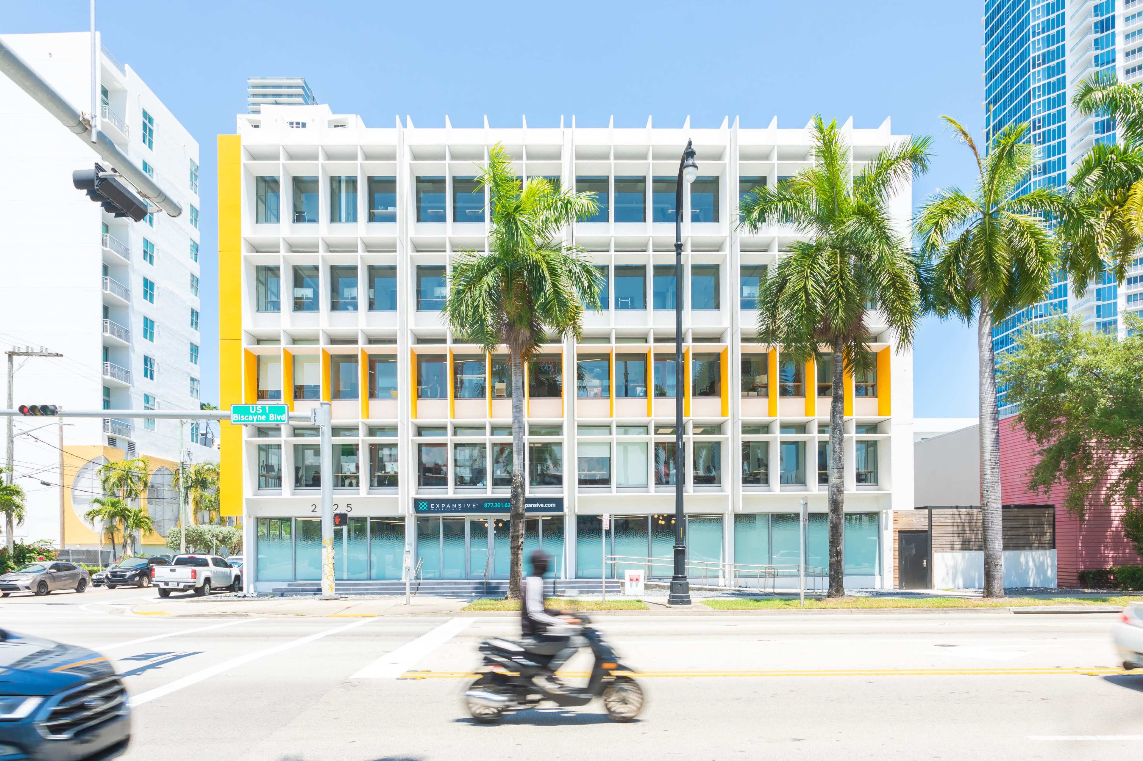 A modern, four-story building with a bright yellow accent and multiple large windows is situated on a busy street lined with palm trees.