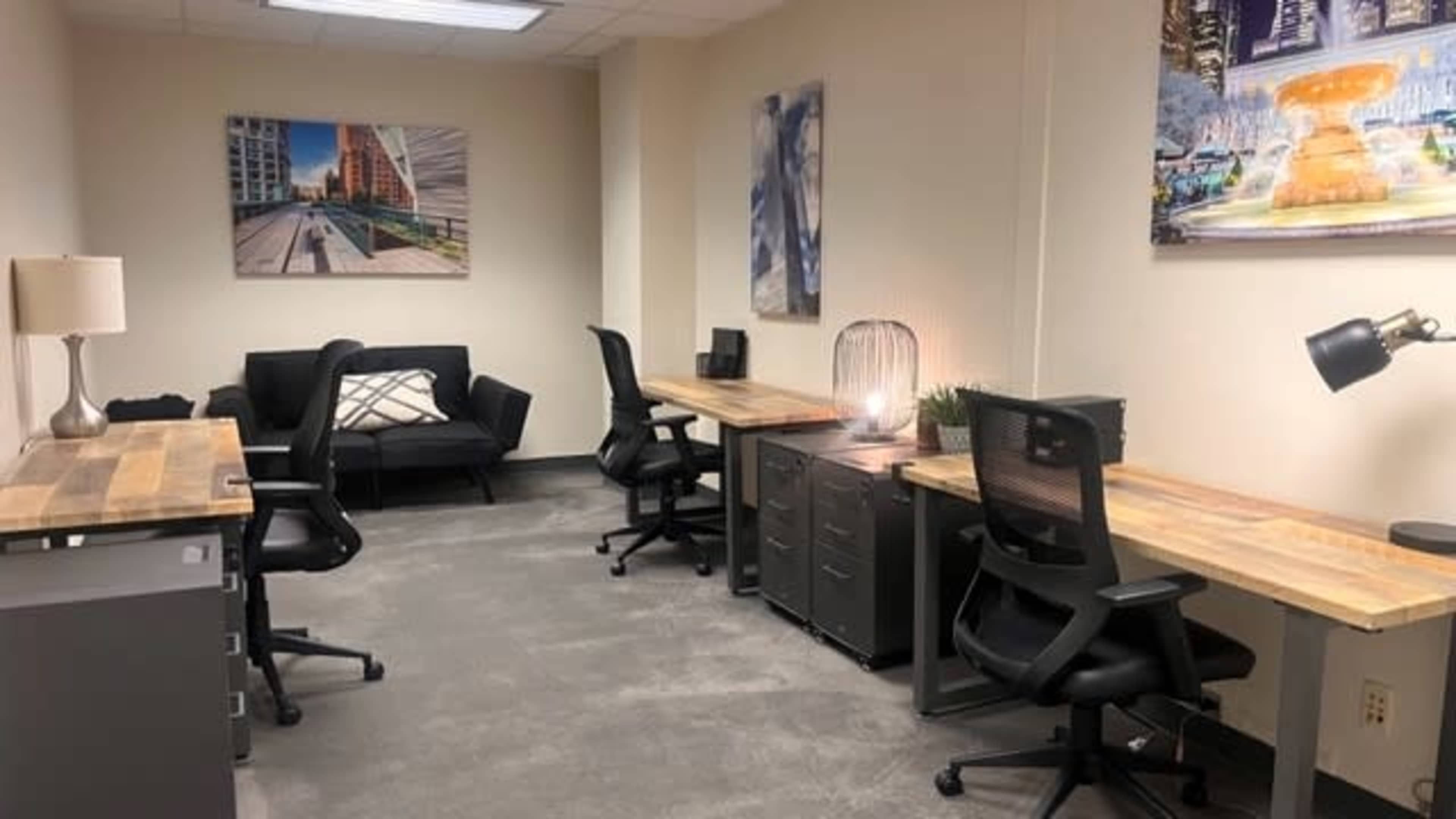 A modern office space featuring two wooden desks with chairs, a couch, and decorative wall art.