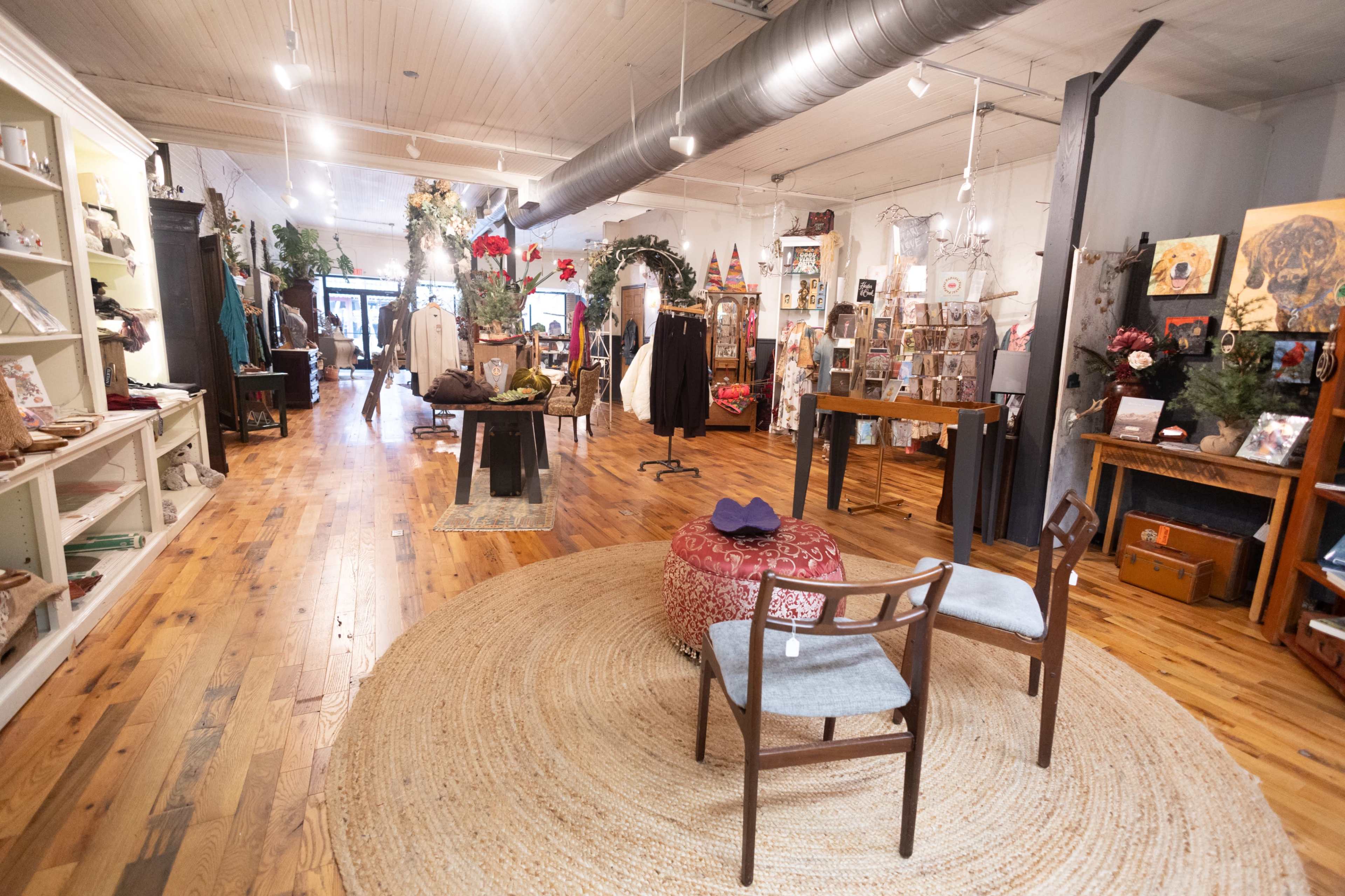 The image shows the interior of a boutique featuring wooden floors, various handmade items displayed on shelves, and an area with two chairs and a round rug.