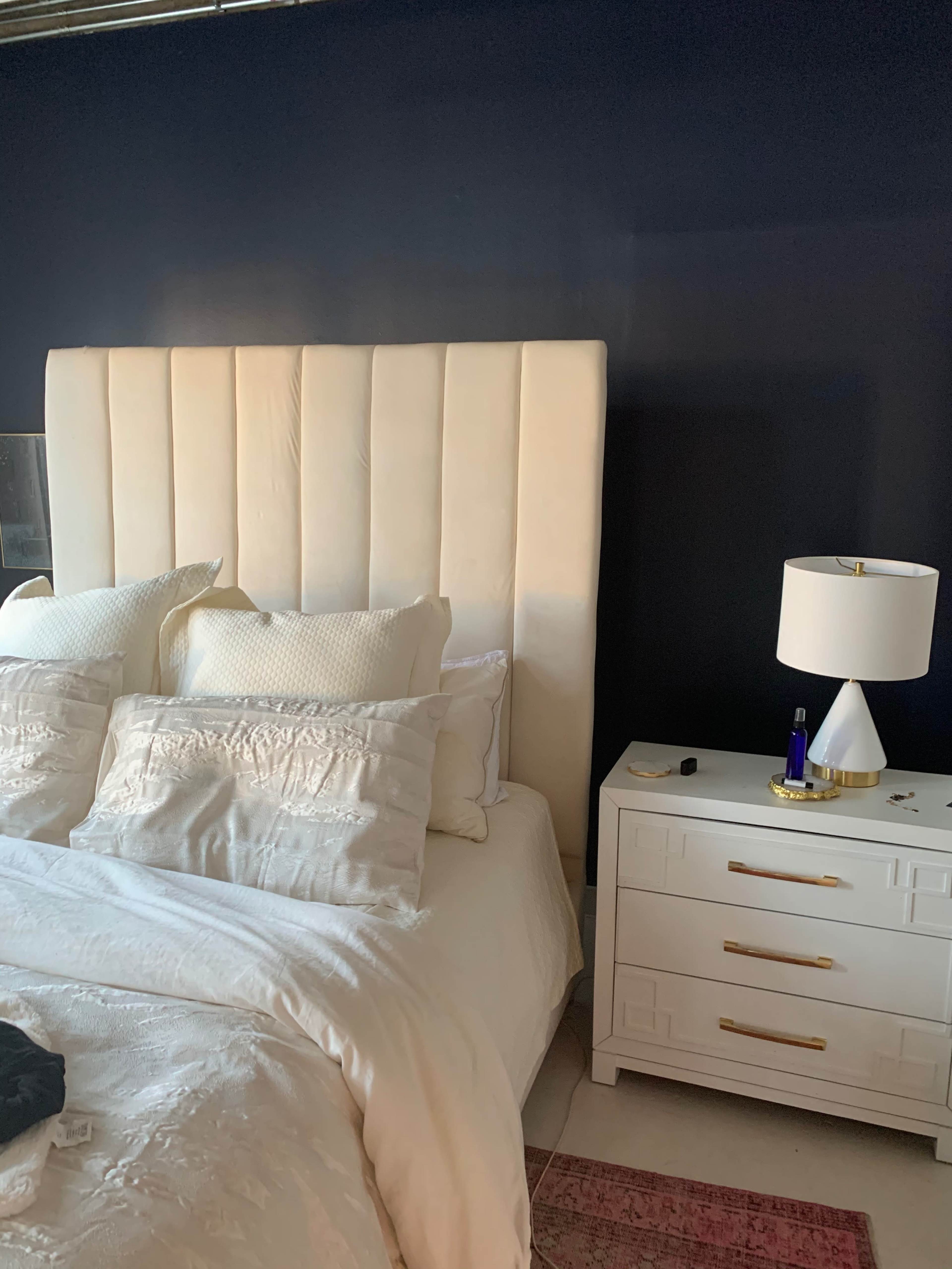 A neatly made bed with white bedding and decorative pillows, accompanied by a white nightstand featuring a lamp, next to a navy blue wall.