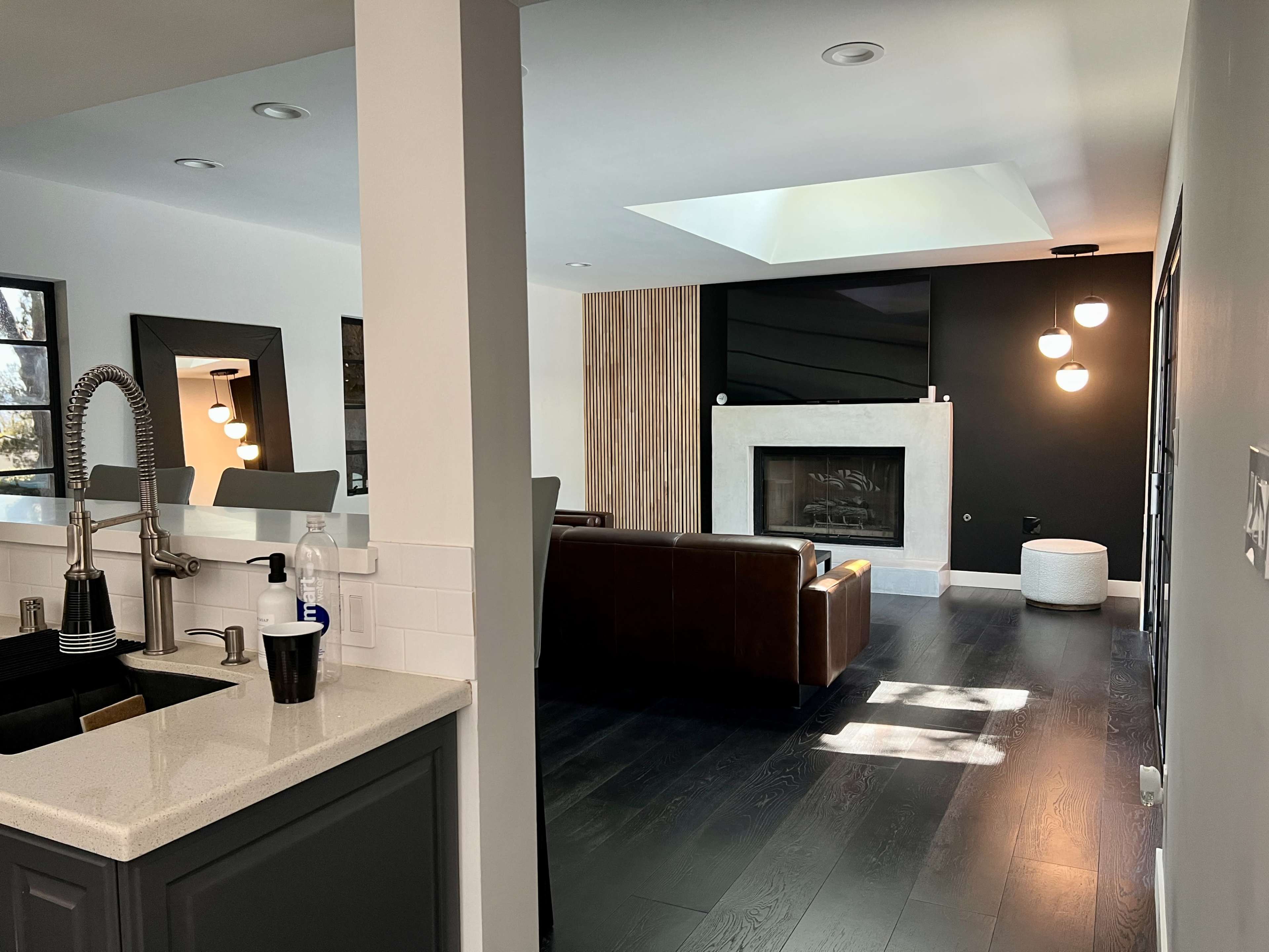 The image shows a modern interior space with a kitchen area in the foreground and a living room featuring a fireplace and large windows in the background.