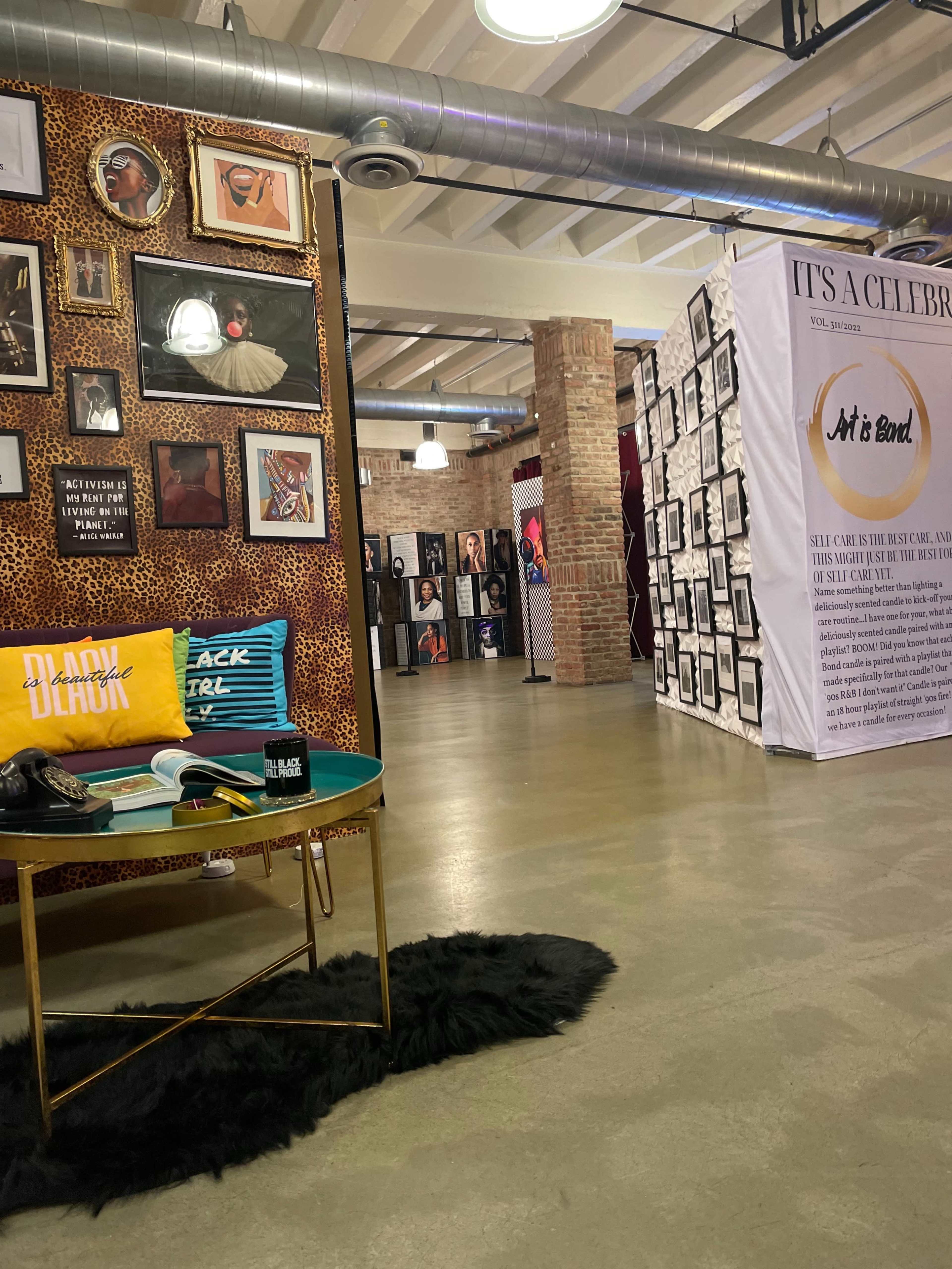 An interior space featuring a leopard print wall with framed portraits, a yellow accent pillow labeled "BLACK" on a table, and an exhibition area displaying various artworks and photographs.