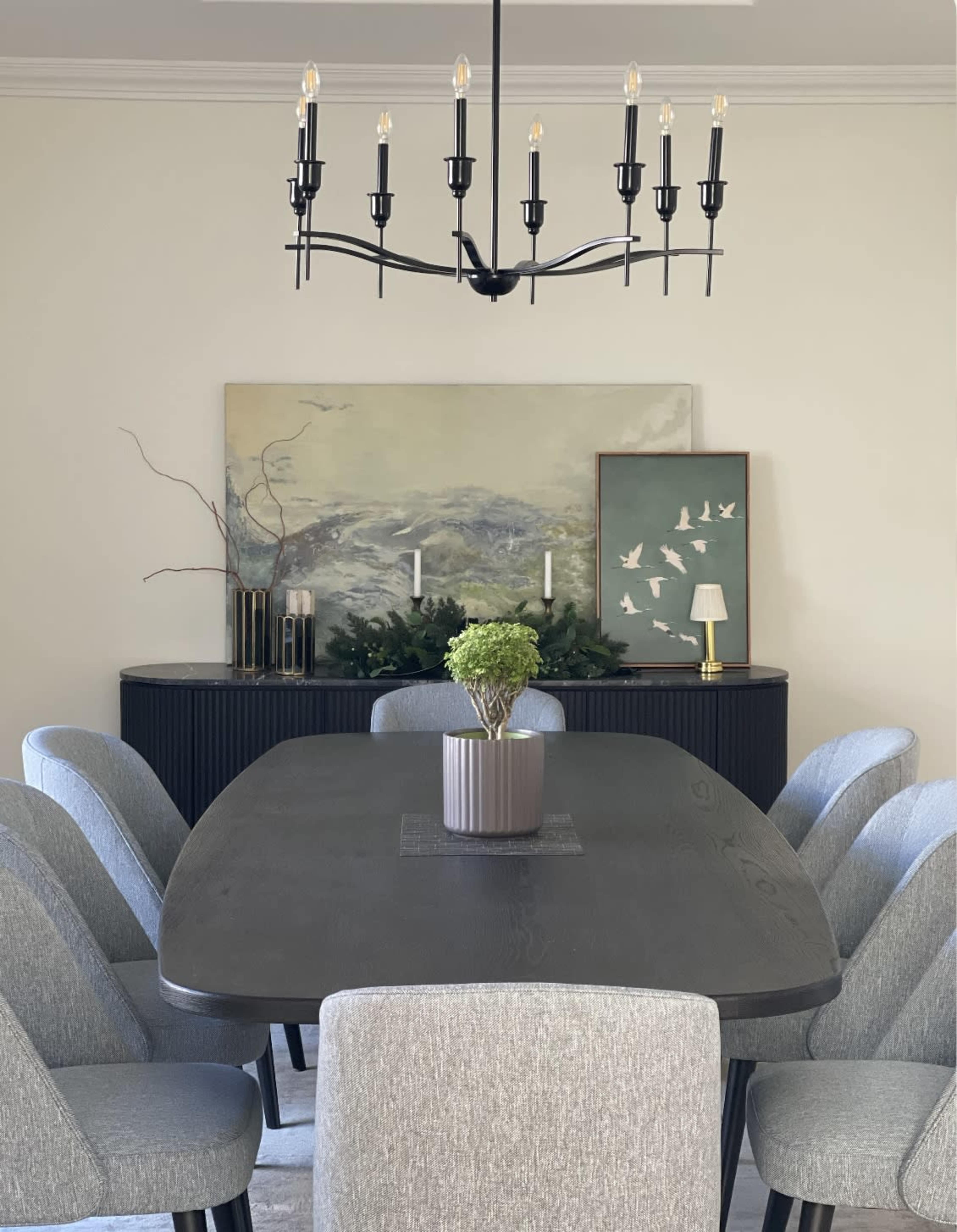 A modern dining room features a large dark table surrounded by gray chairs, with a chandelier overhead and a sideboard displaying decorative items and a landscape painting.