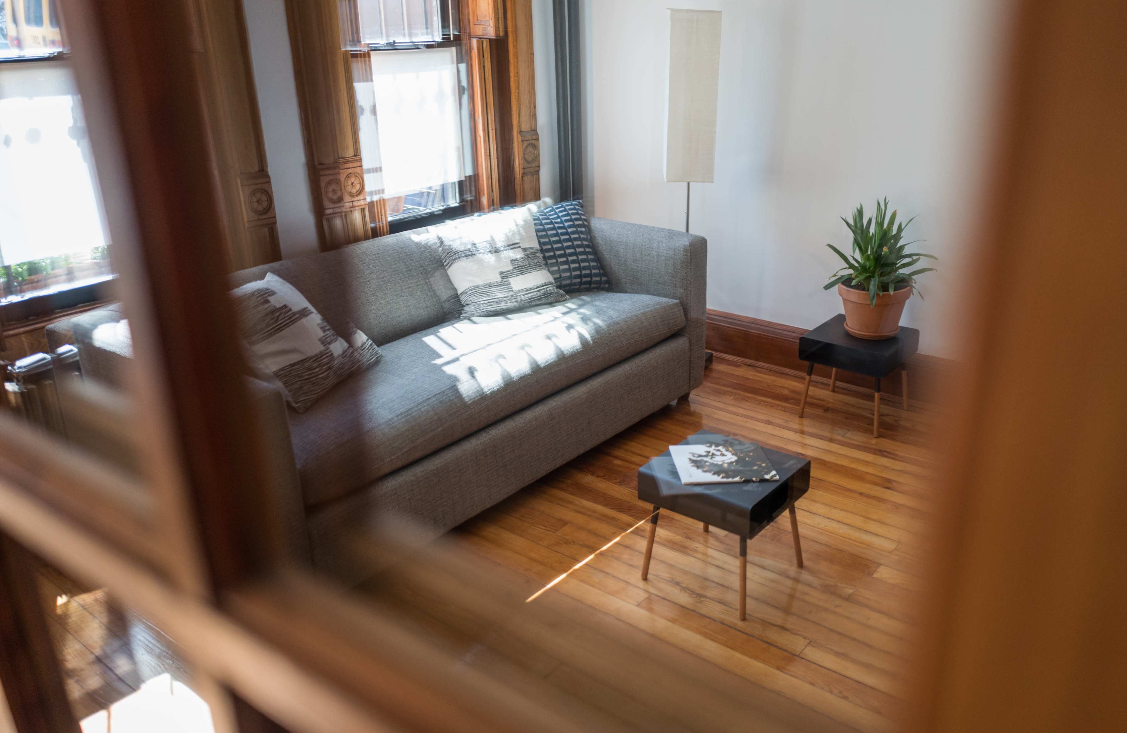 A gray sofa with decorative pillows sits in a room with wooden floors, next to a small black table and a potted plant.
