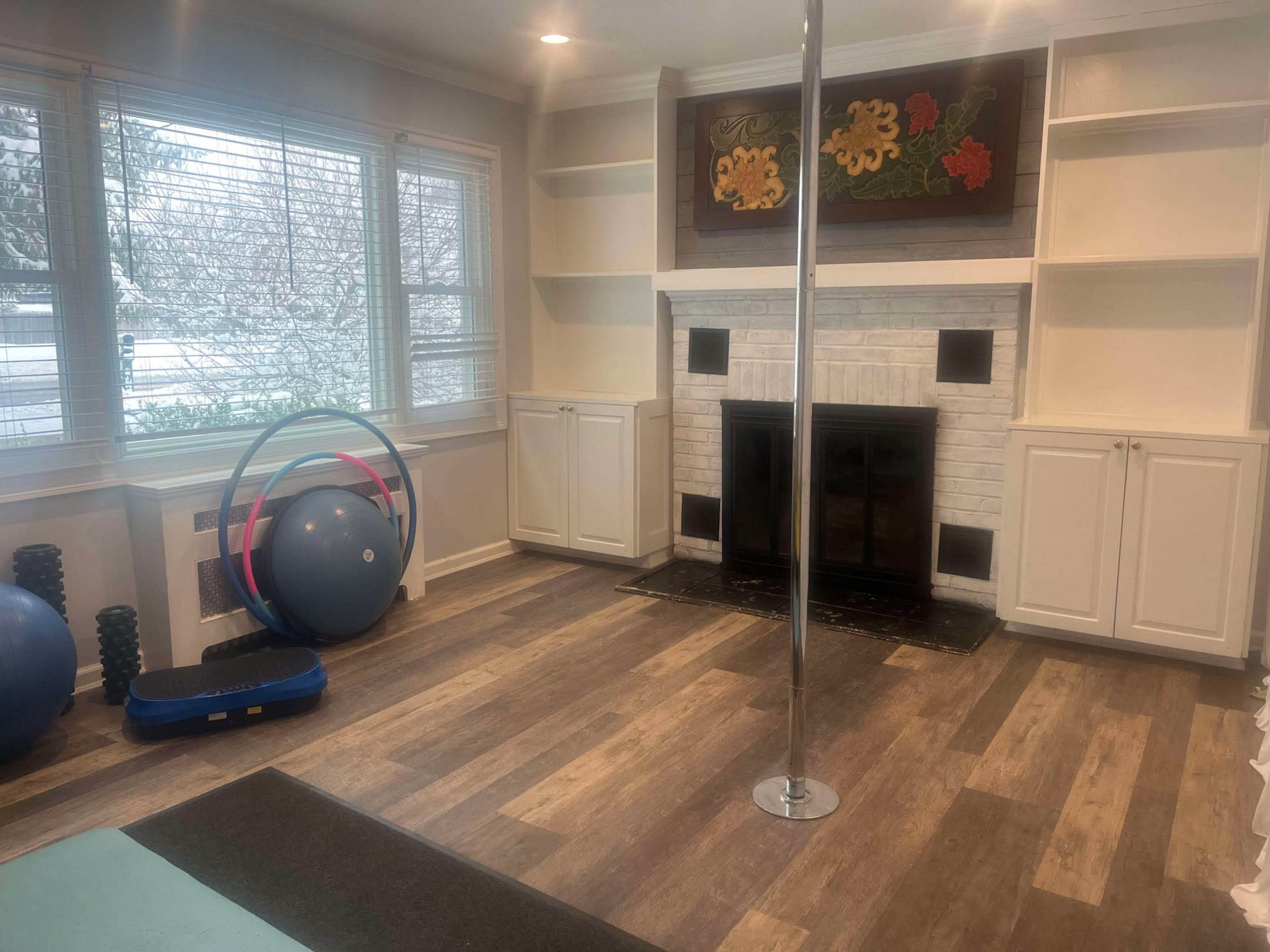 The image shows a home exercise room featuring a pole, a large exercise ball, a step platform, and a fireplace with built-in shelving.