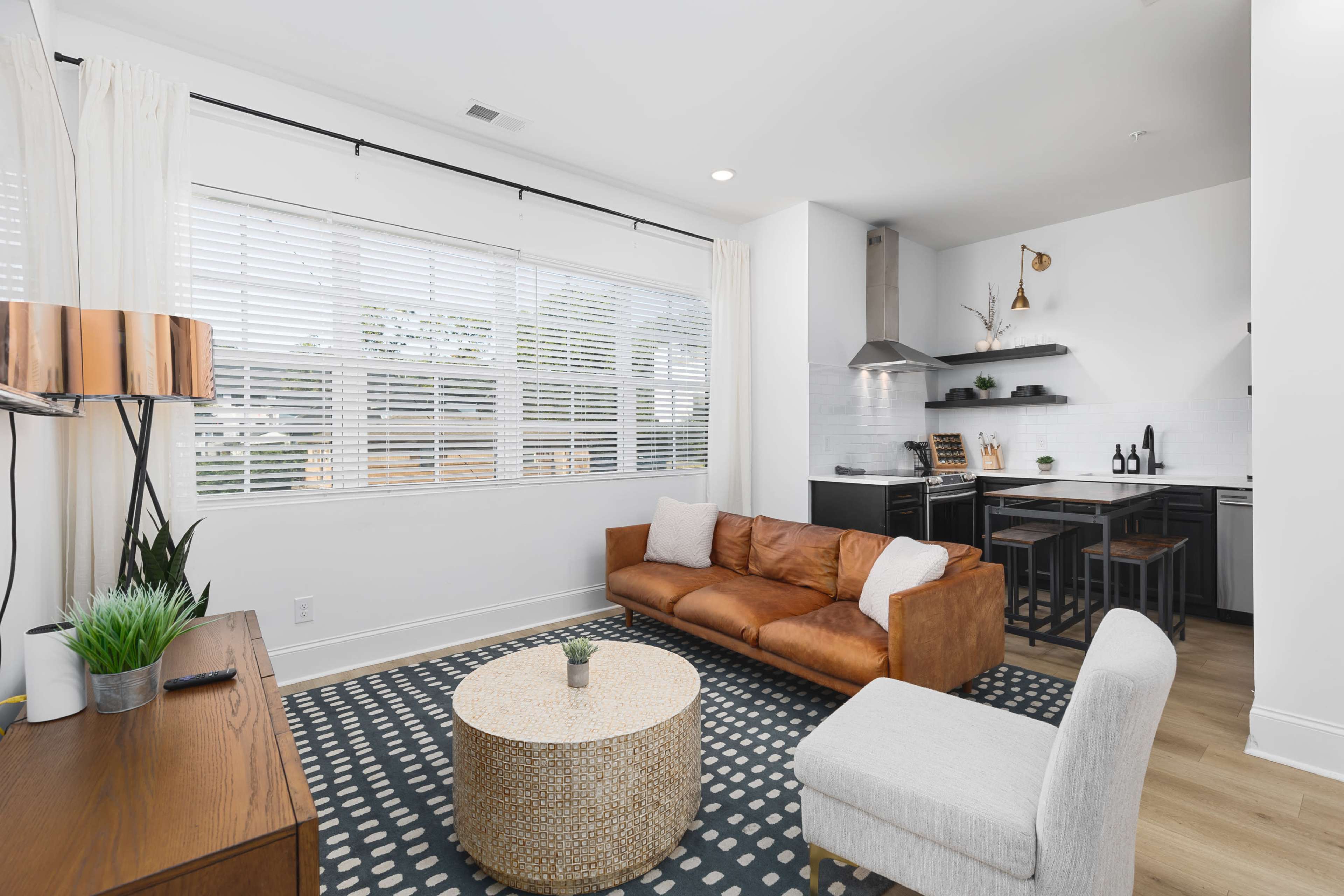 A modern living room features a brown leather sofa, a round coffee table, and a kitchen area with black cabinetry and stainless steel appliances.