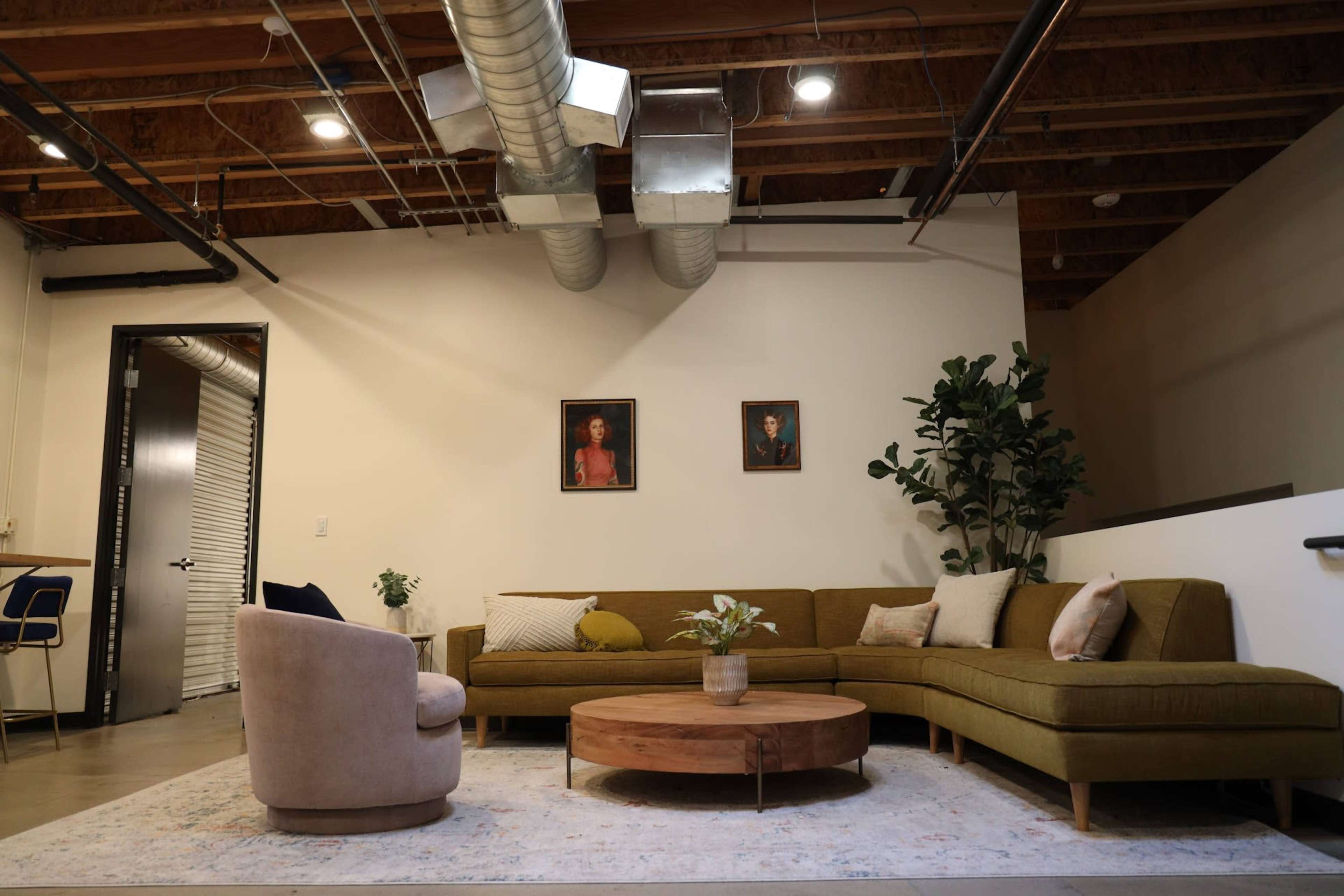 The image shows a modern lounge area featuring a green sectional sofa, a round wooden coffee table, and two framed portraits on the wall.
