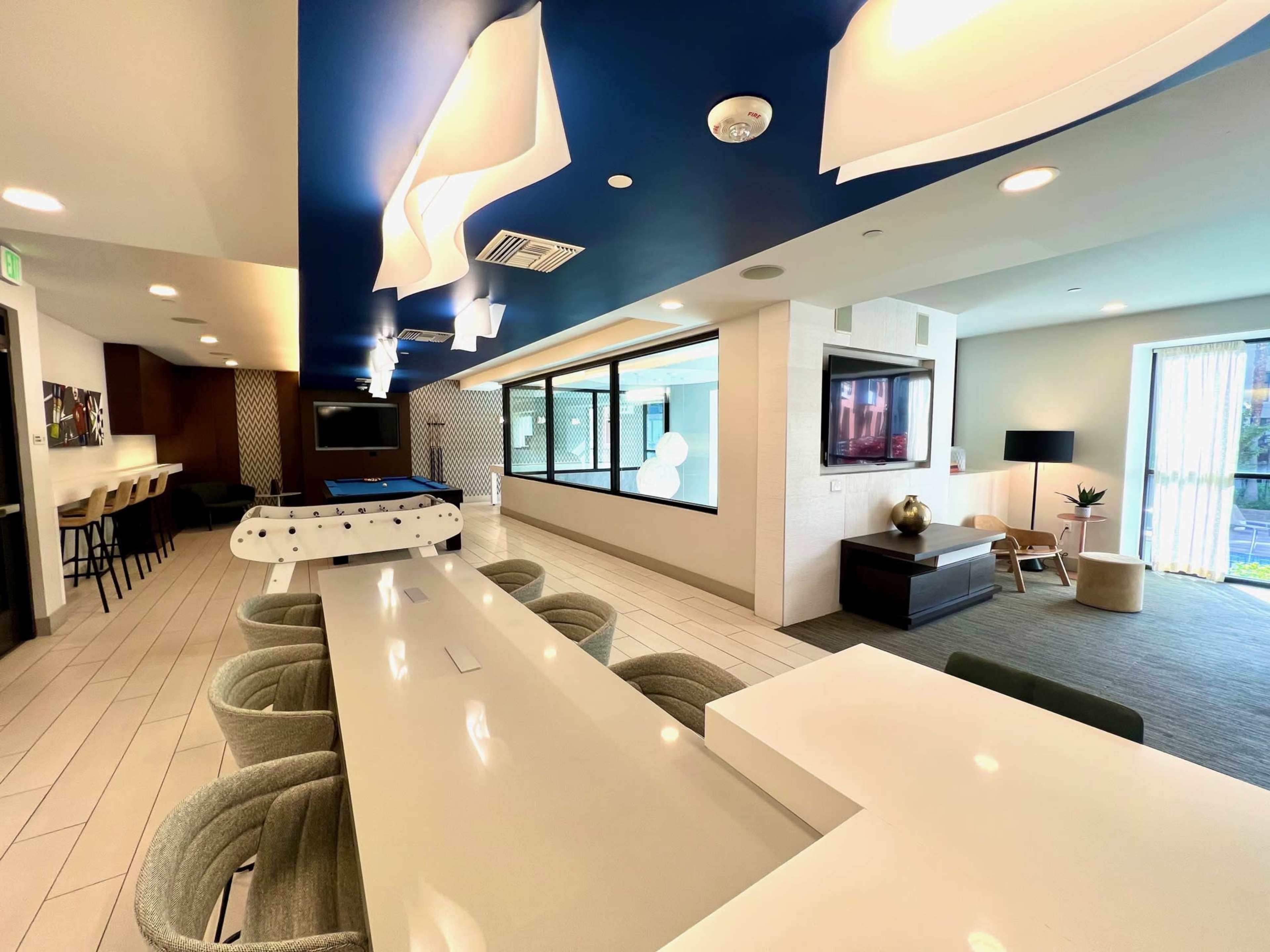 A modern clubhouse area featuring a long white table, bar seating, a pool table, and large windows overlooking a lounge space.