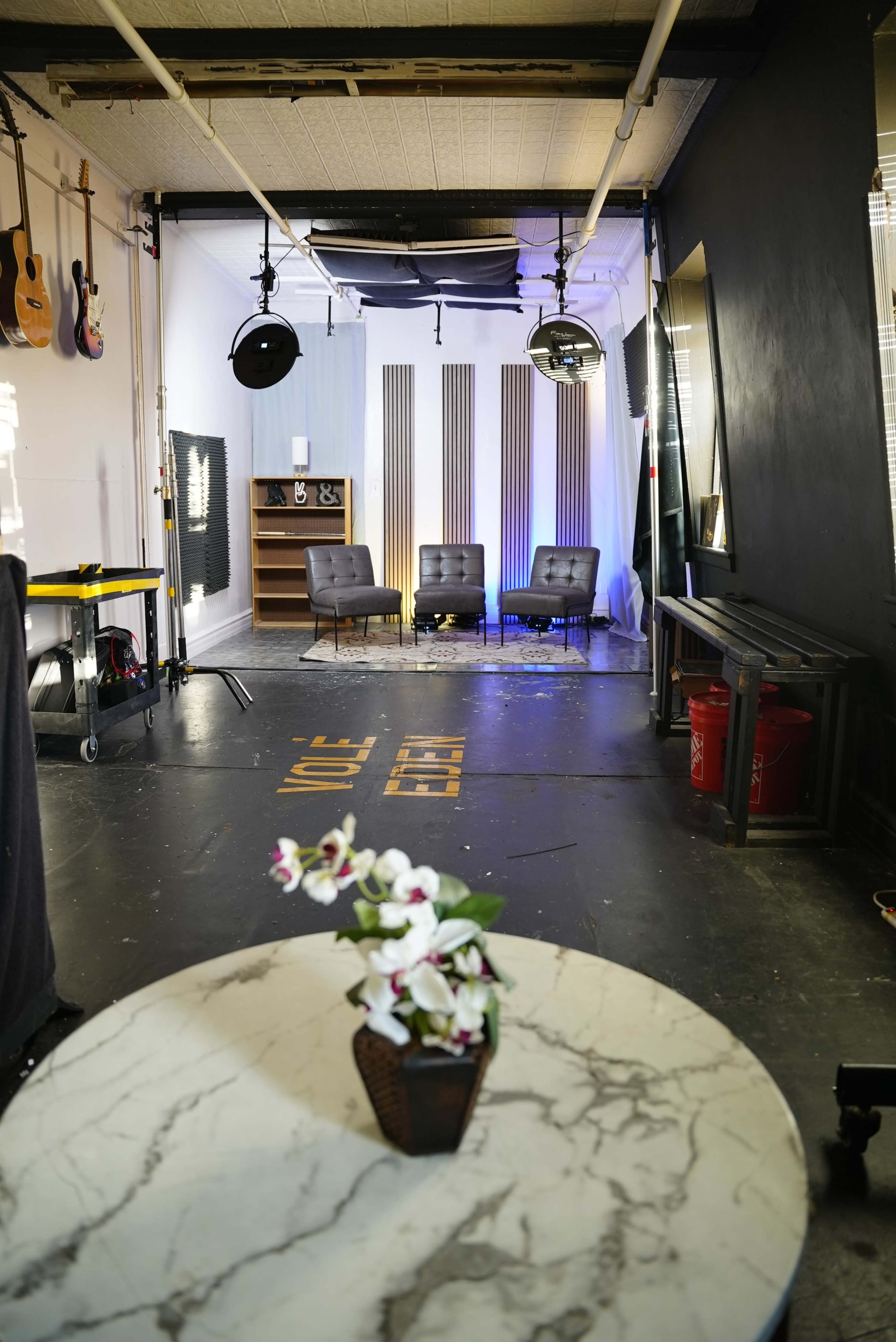 The image shows a well-lit studio space with three black chairs, a marble coffee table with a flower arrangement, and various lighting and sound equipment in the background.