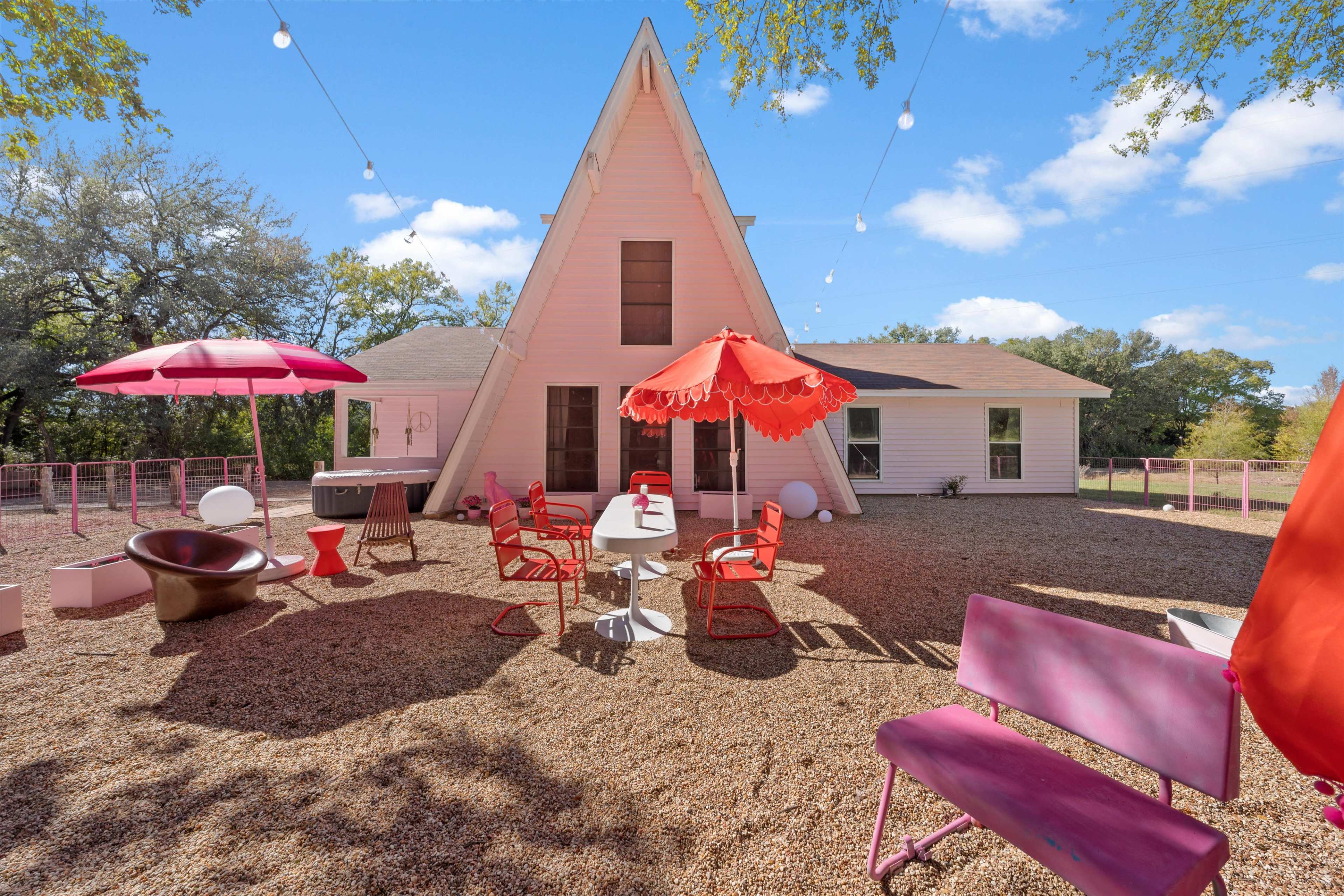 A modern A-frame house with a gravel patio features colorful outdoor furniture and decorative lighting strung above.