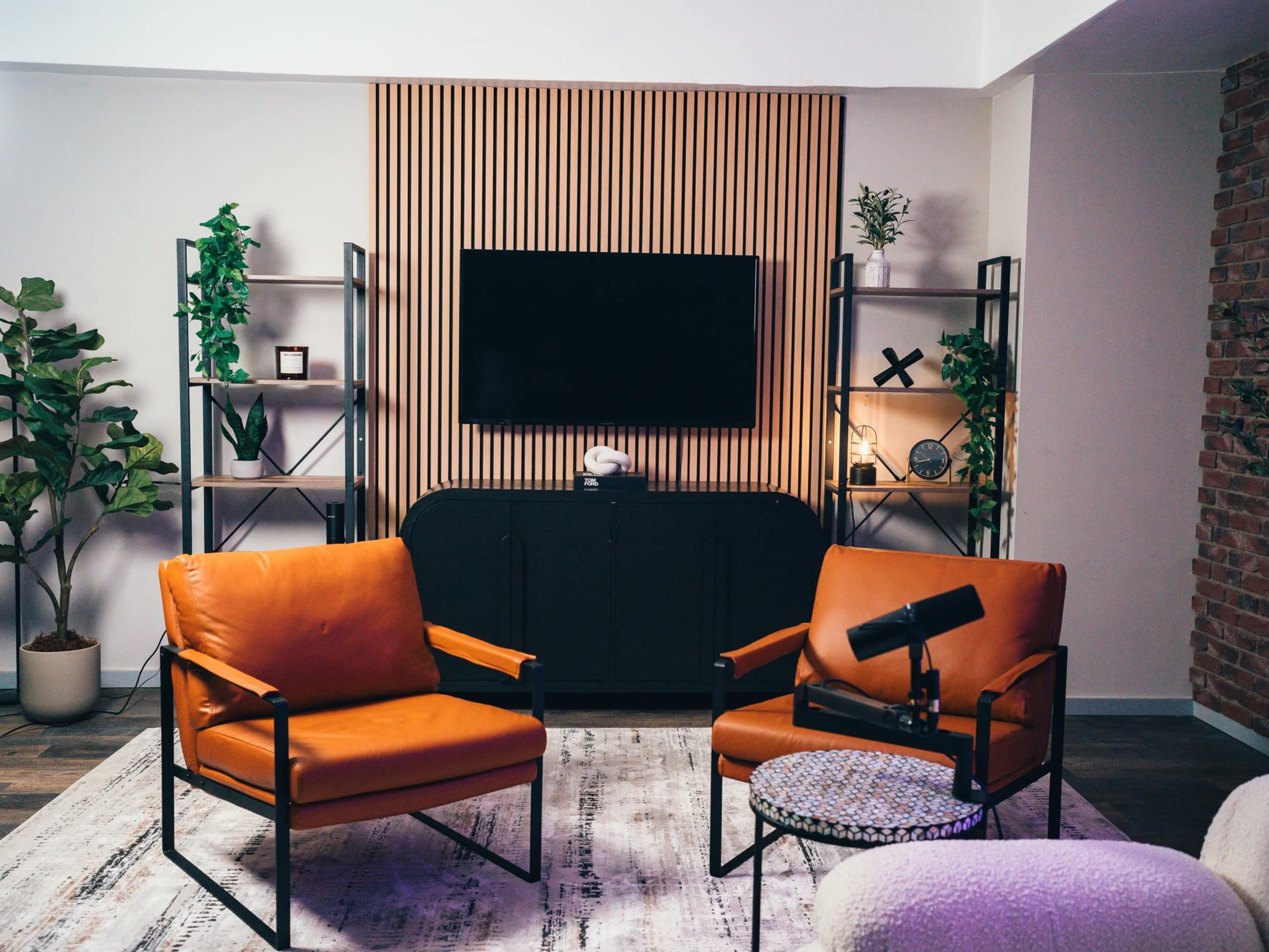 A modern living room features two orange chairs, a black cabinet, a wall-mounted TV, and decorative shelving with plants.