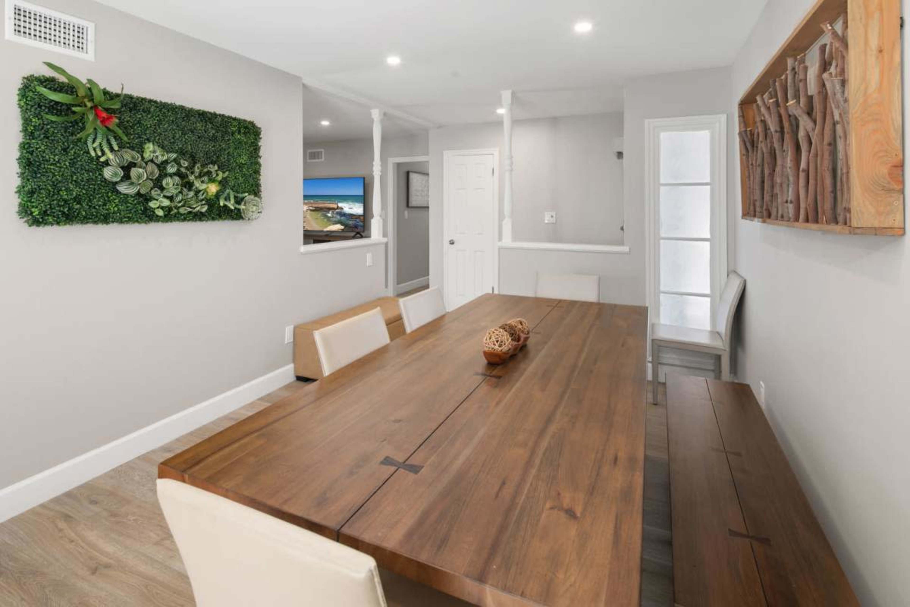 The image shows a modern dining room with a wooden table surrounded by chairs, featuring a green wall decoration and a door leading to another room.