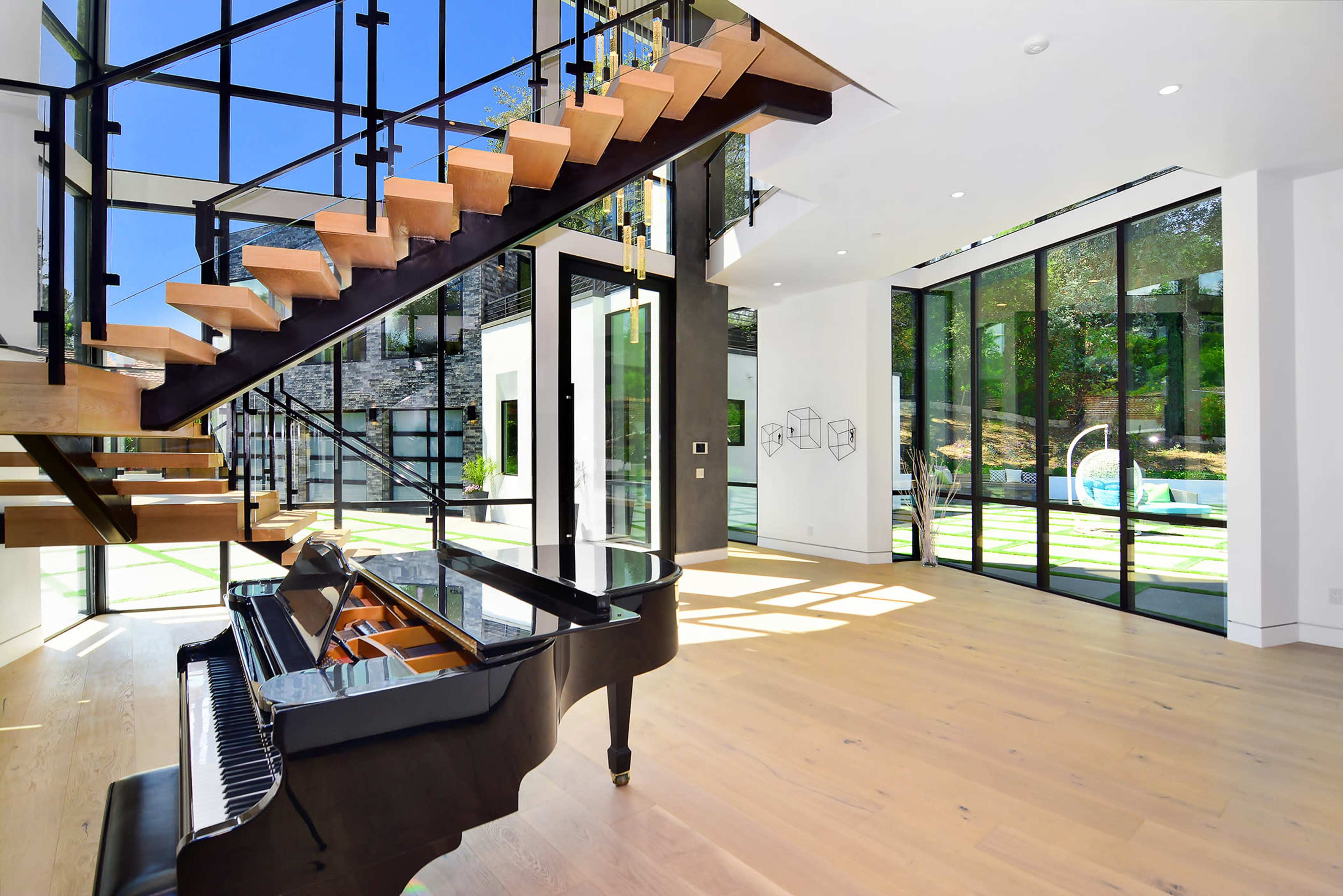 A modern open-concept living space with a grand piano positioned near a staircase and large glass doors that lead to an outdoor area.
