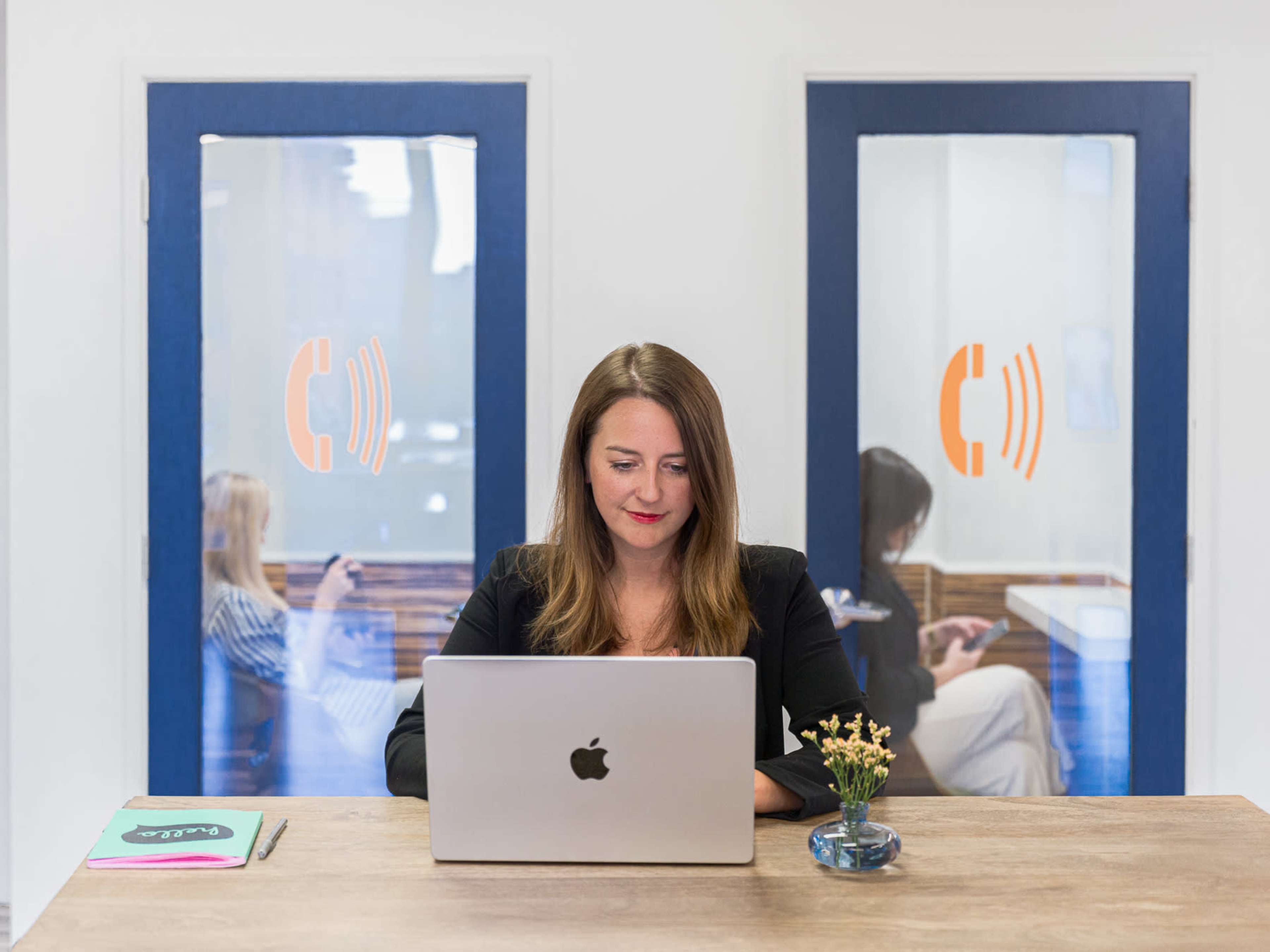 A woman sits at a wooden desk using a laptop, with two people visible through glass doors in the background.