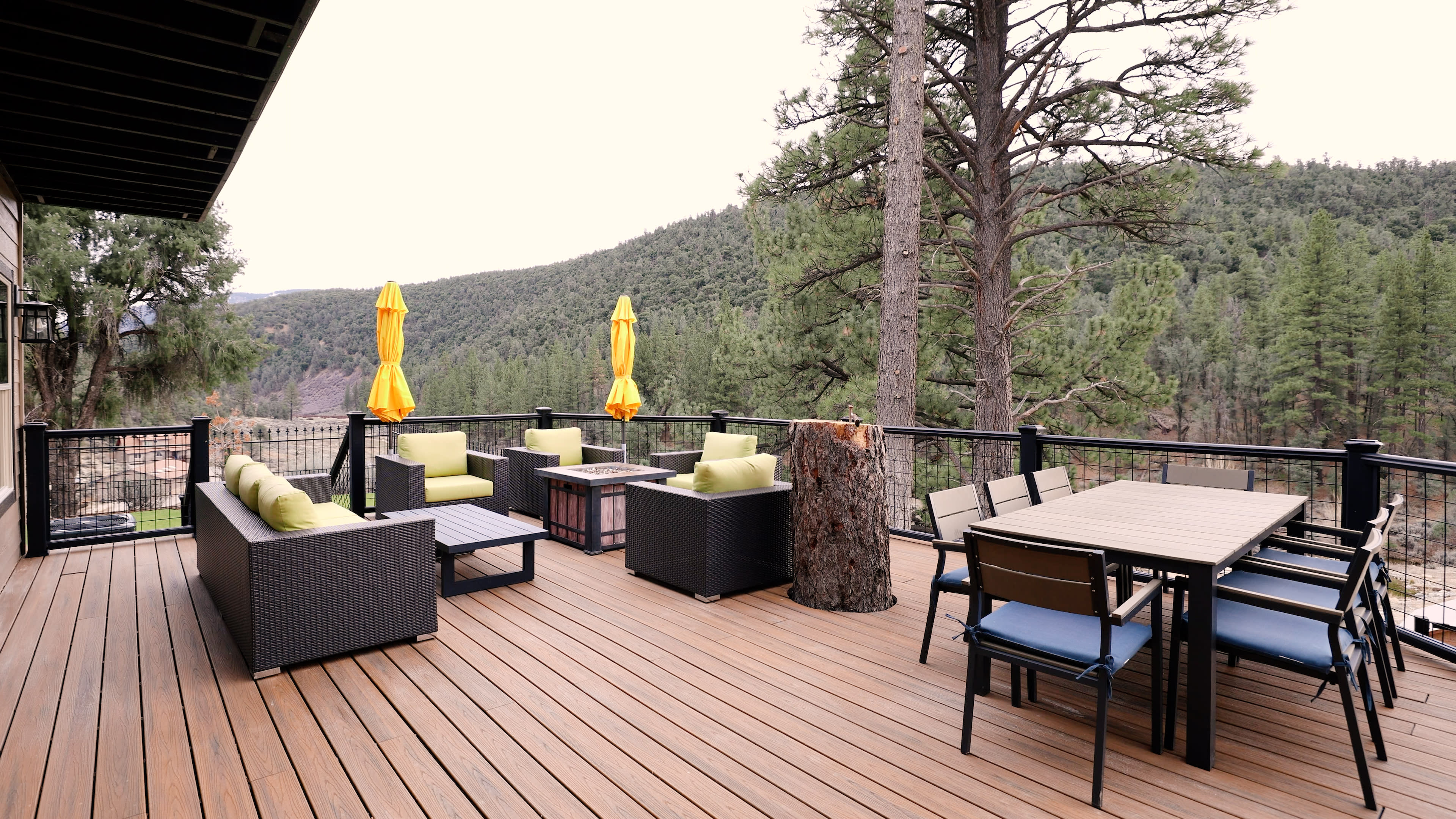 The image shows a spacious wooden deck adorned with outdoor furniture, including seating areas, a dining table, and yellow umbrellas, set against a backdrop of a forested hillside.