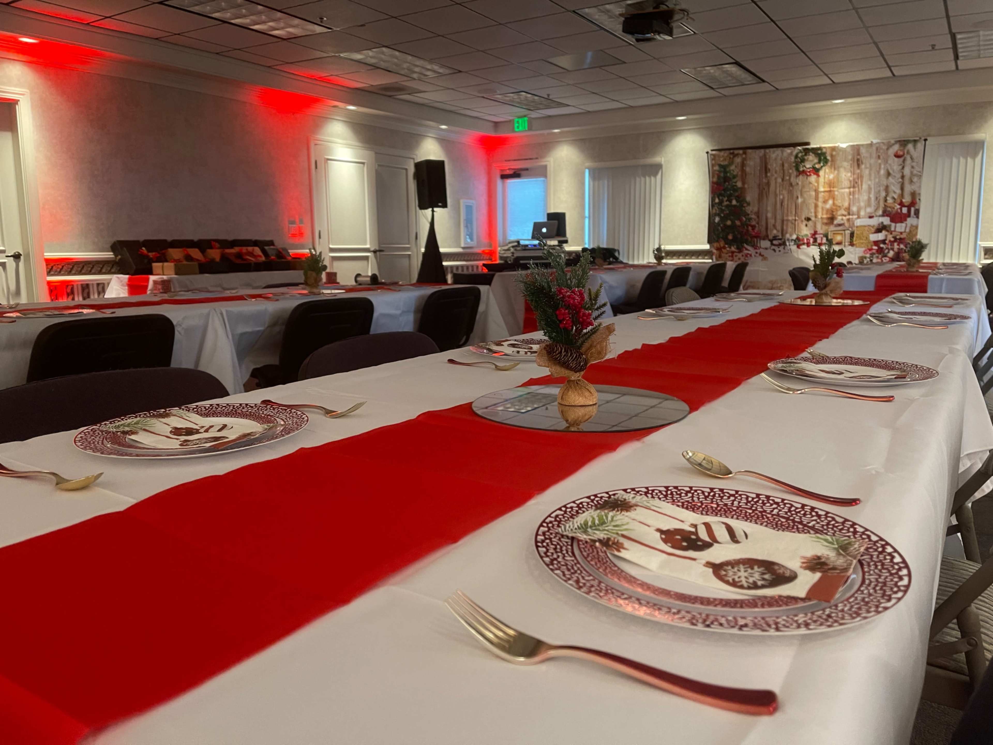 A long table set for an event features red table runners, decorative plates, and small floral centerpieces in a well-lit room with soft red lighting.