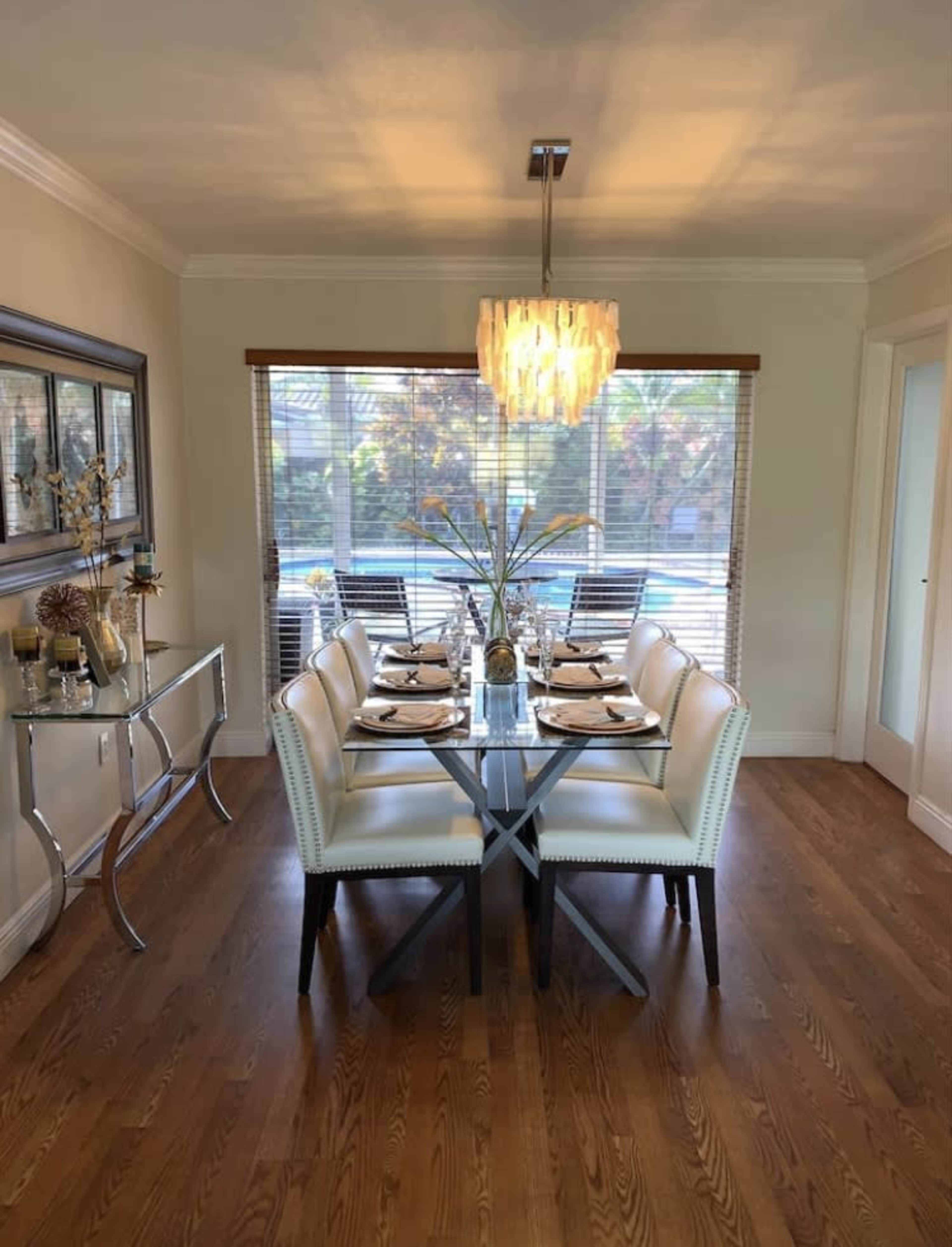 A modern dining room features a long glass table surrounded by white upholstered chairs, illuminated by an elegant chandelier and natural light from large windows.