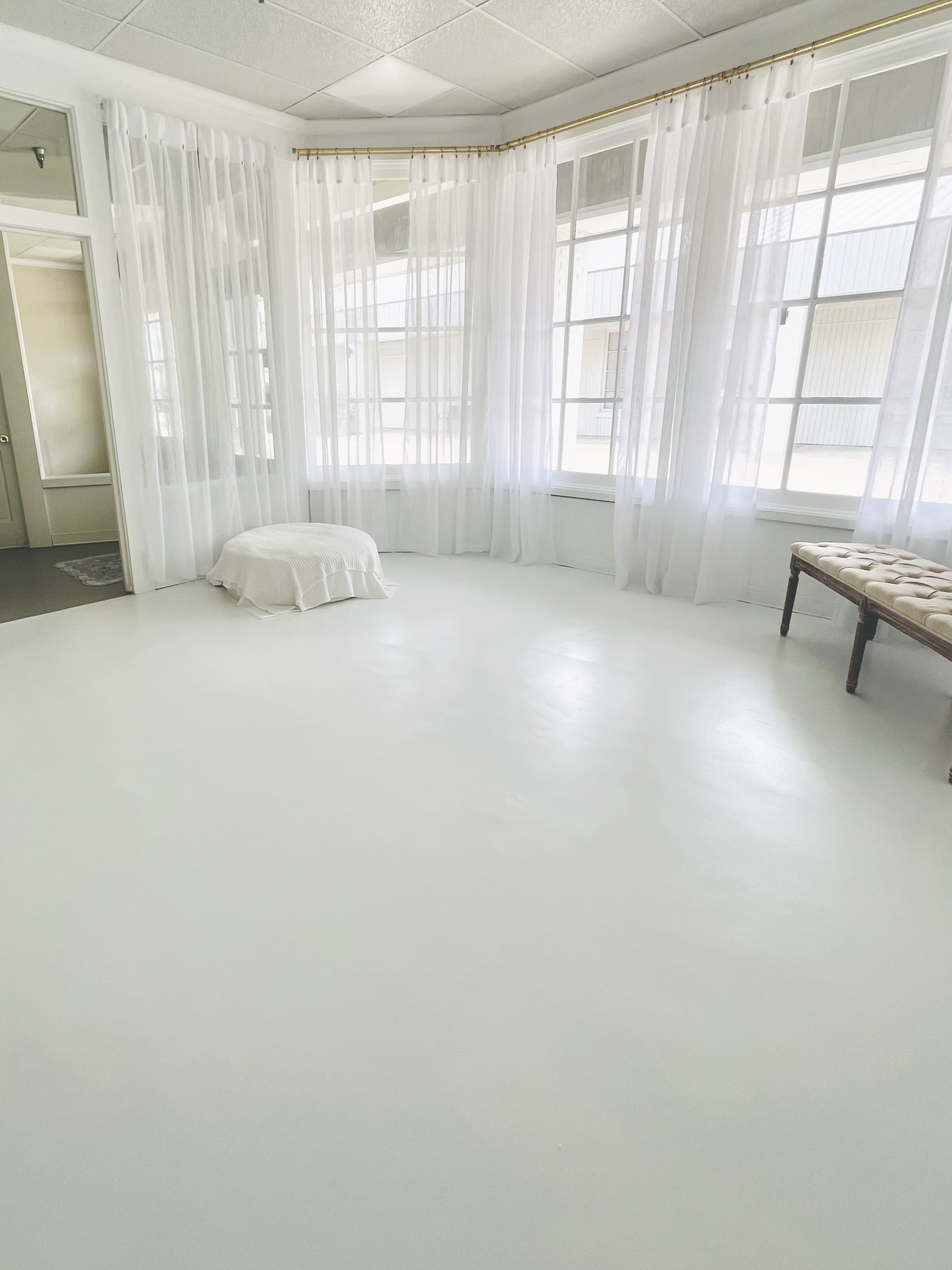 The image shows a bright, empty room with large windows draped in sheer white curtains and a small white bench in the corner.