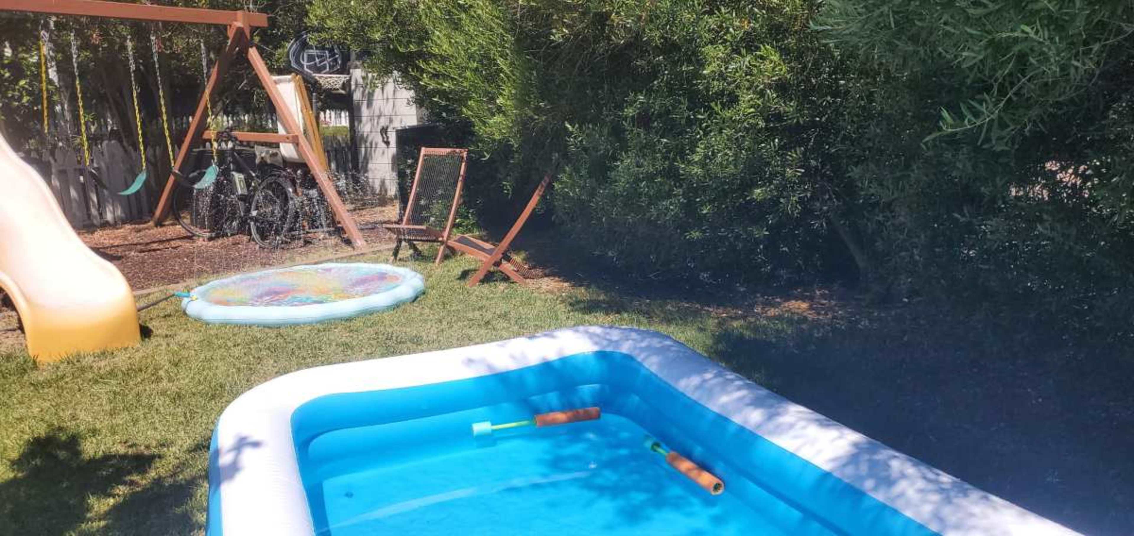 A small inflatable pool sits in a backyard next to a water slide and a playground swing set.