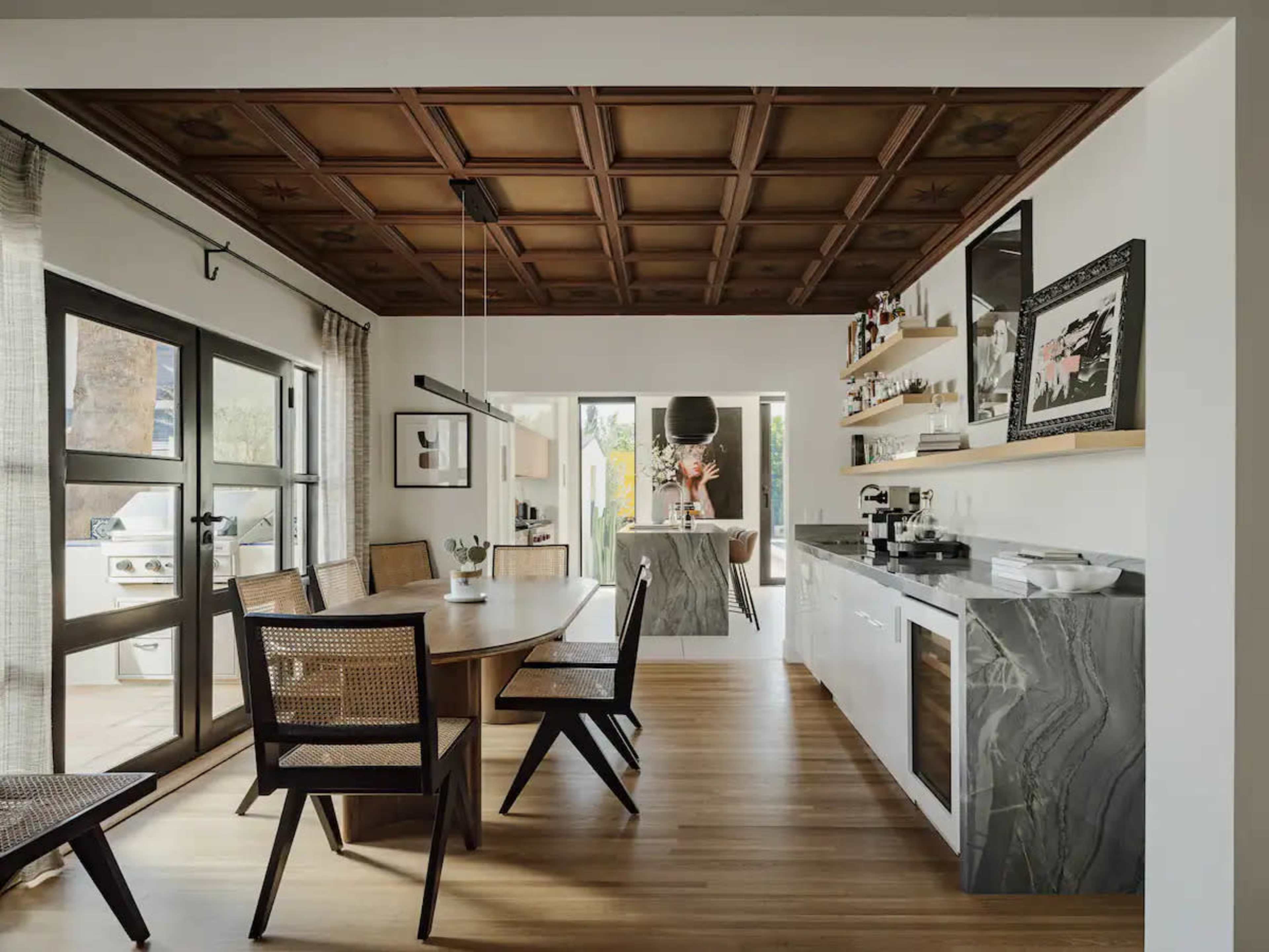 A modern dining area featuring a wooden coffered ceiling, a round table with six chairs, and a sleek kitchen with marble accents and open shelving.