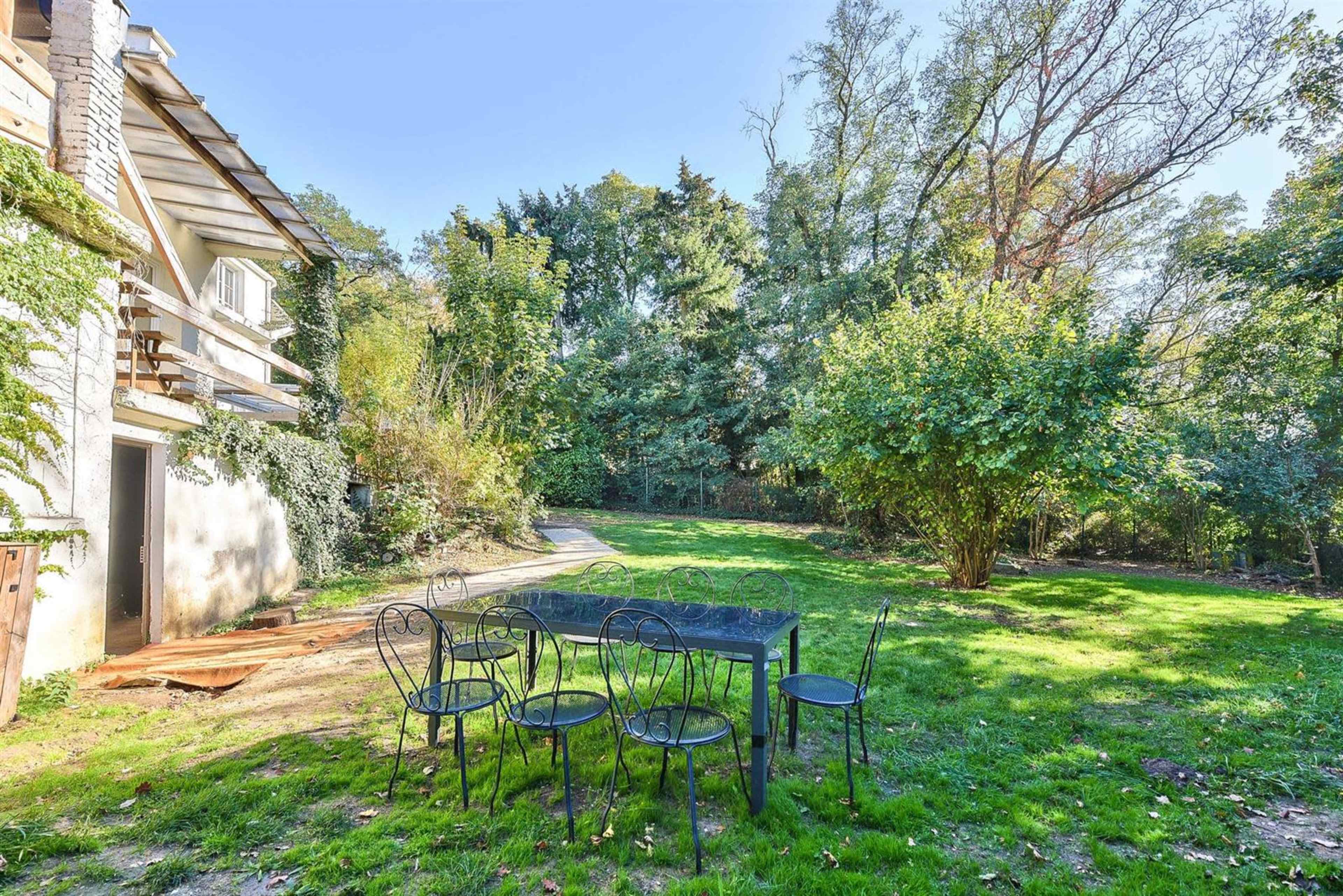 A small black table with six chairs sits on a grassy area beside a house, surrounded by trees and vegetation.