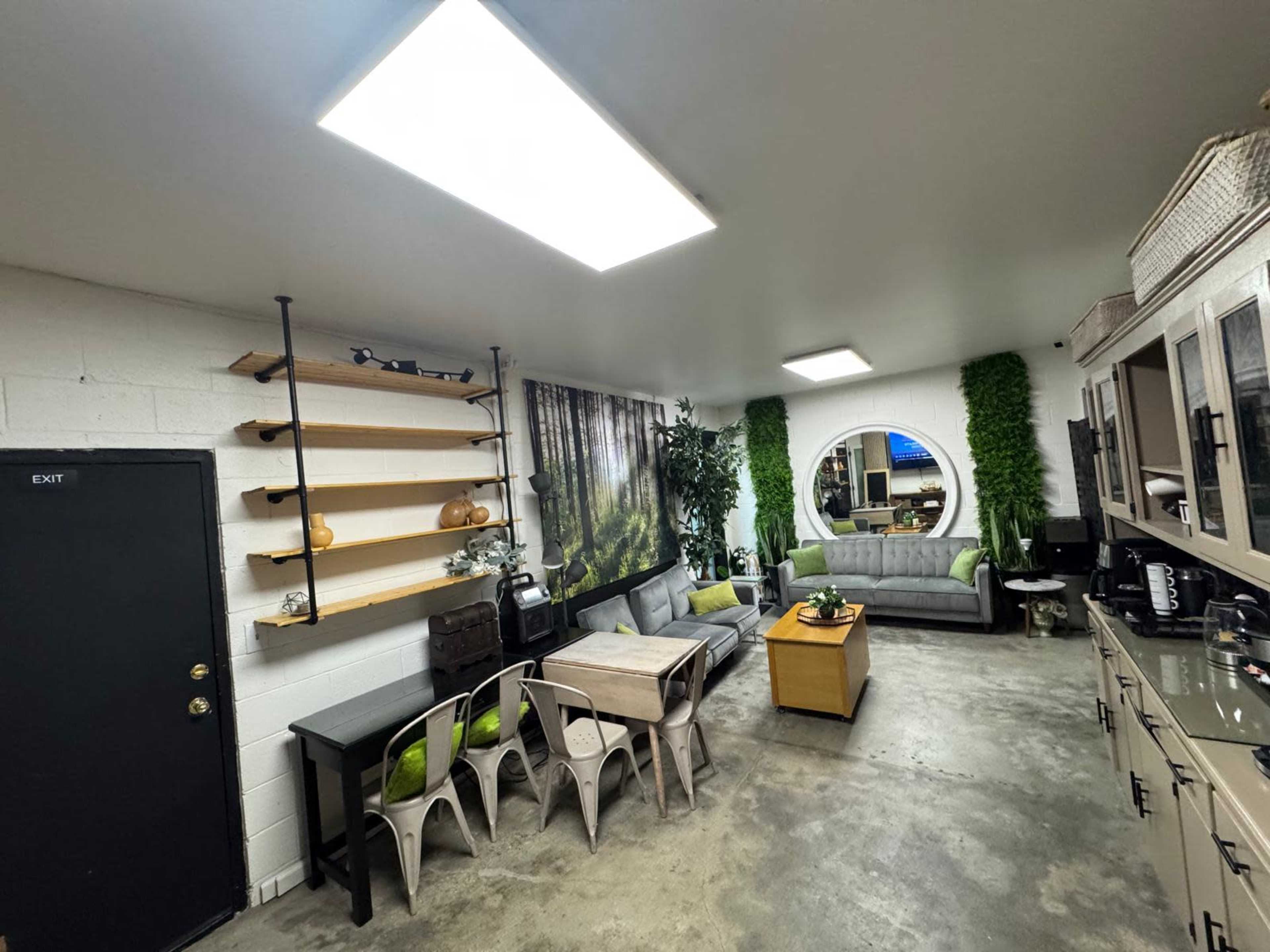 The image shows a modern, open lounge area with a grey sofa, wooden shelves on the walls, and a round mirror opposite the exit door.