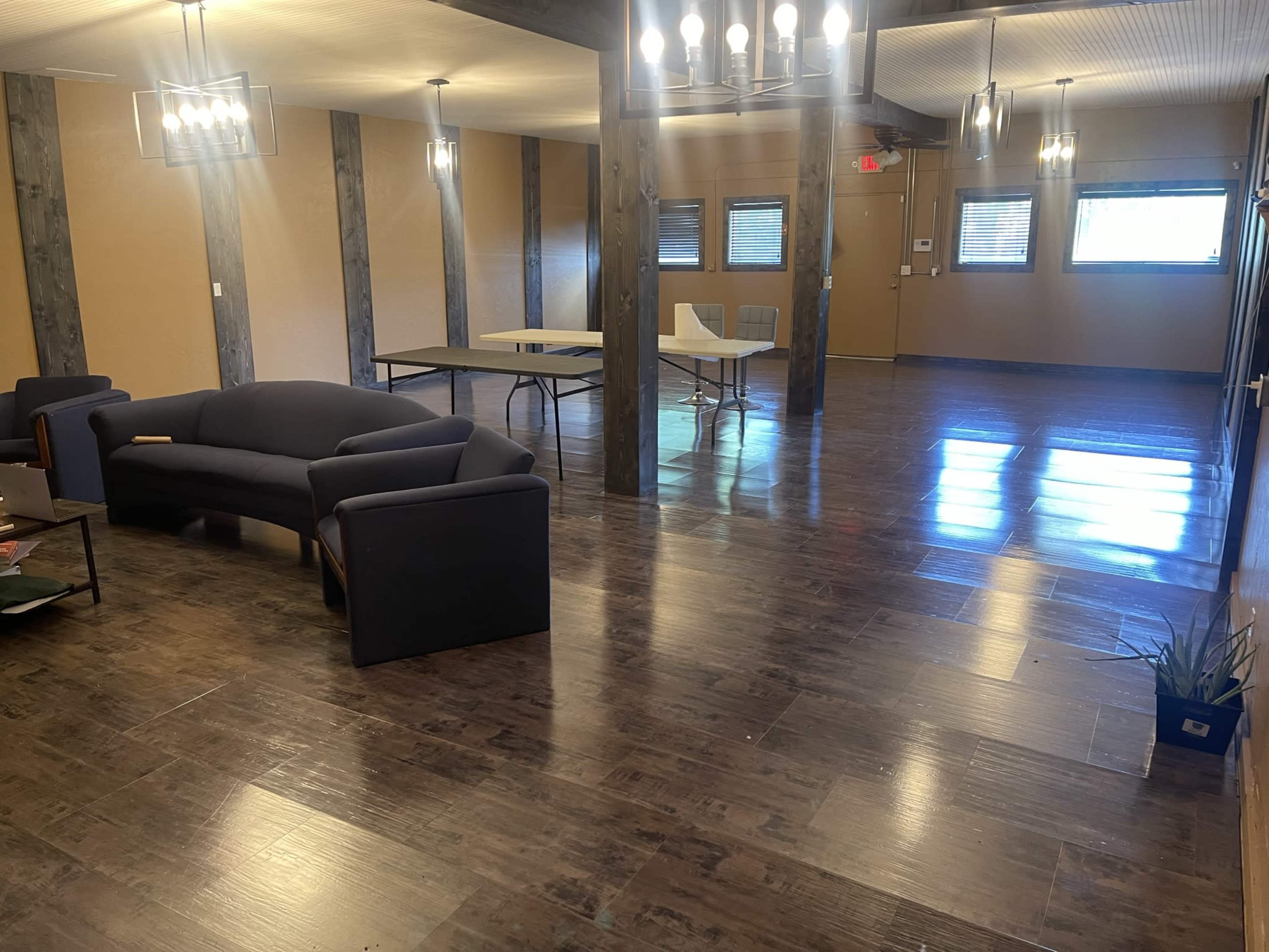 A spacious room with wooden flooring, featuring a blue couch, a few tables, and large windows.