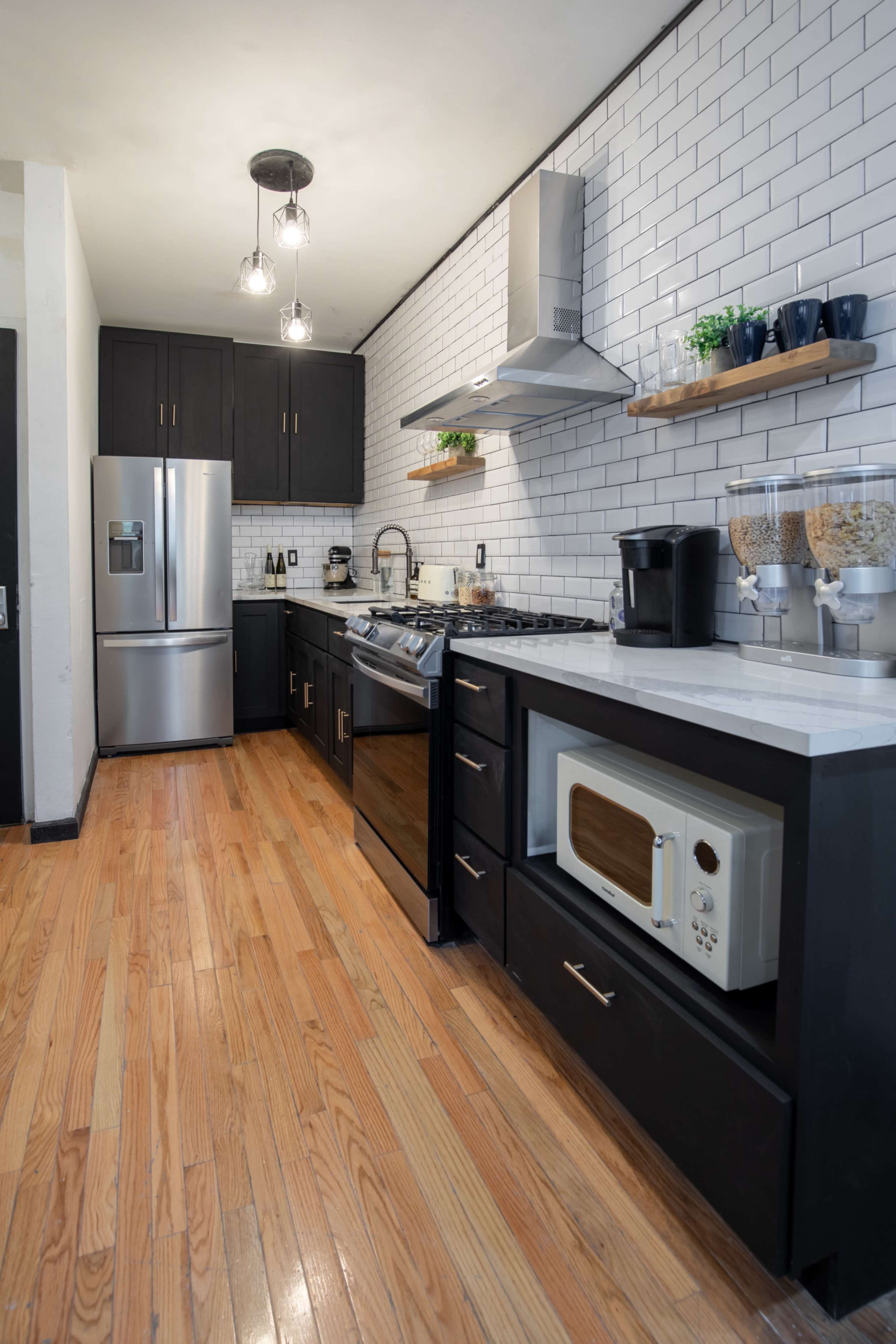 A modern kitchen with stainless steel appliances, black cabinetry, and white subway tile walls, featuring hardwood floors.