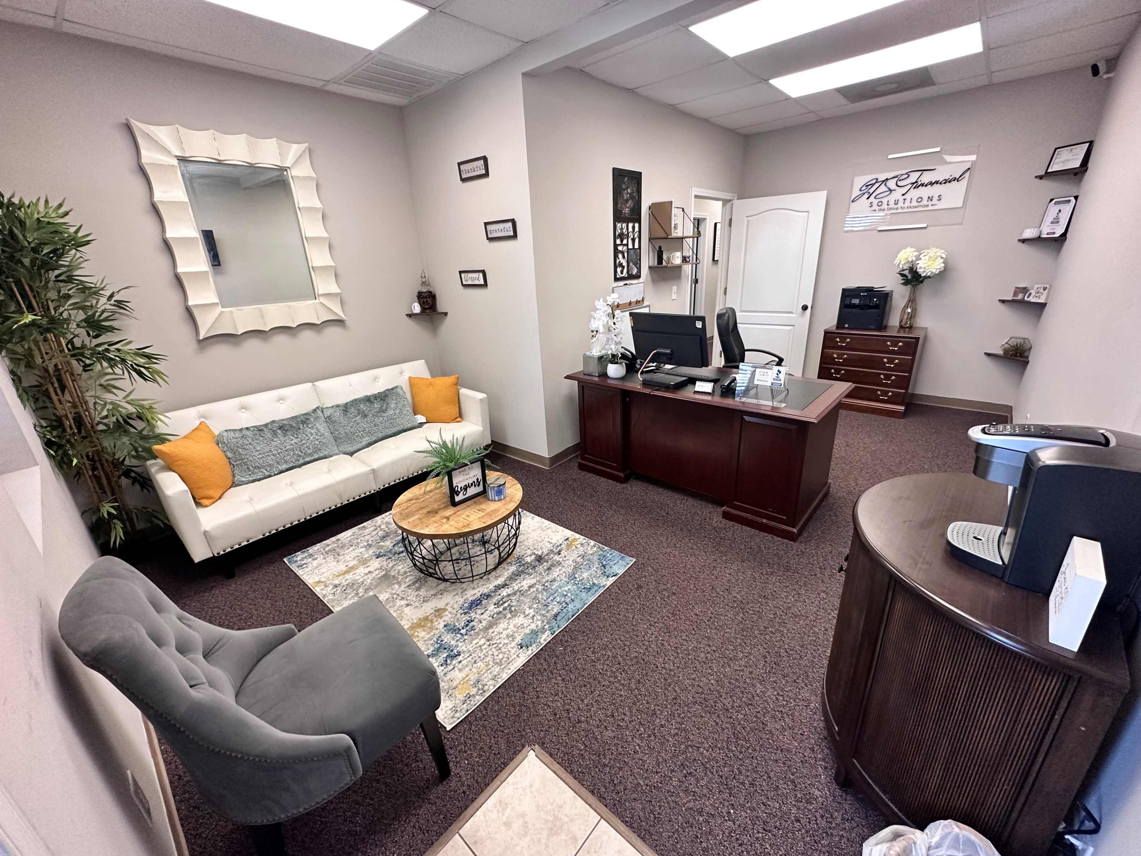 A neatly arranged office with a reception area featuring a white couch, a round coffee table, a desk, and various decorative elements.