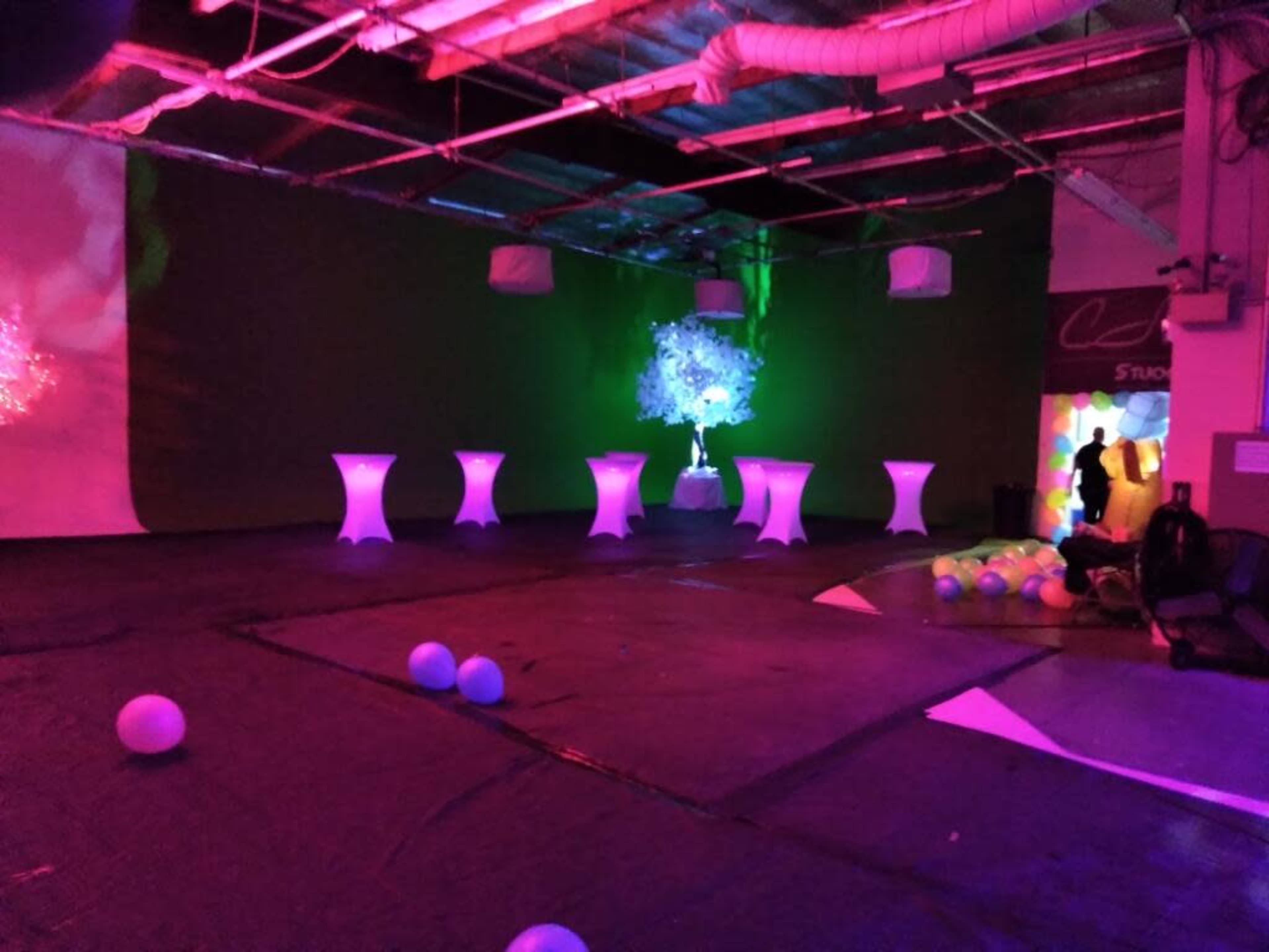 The scene features a dimly lit event space with illuminated tables, a decorative tree, and scattered balloons on the floor.