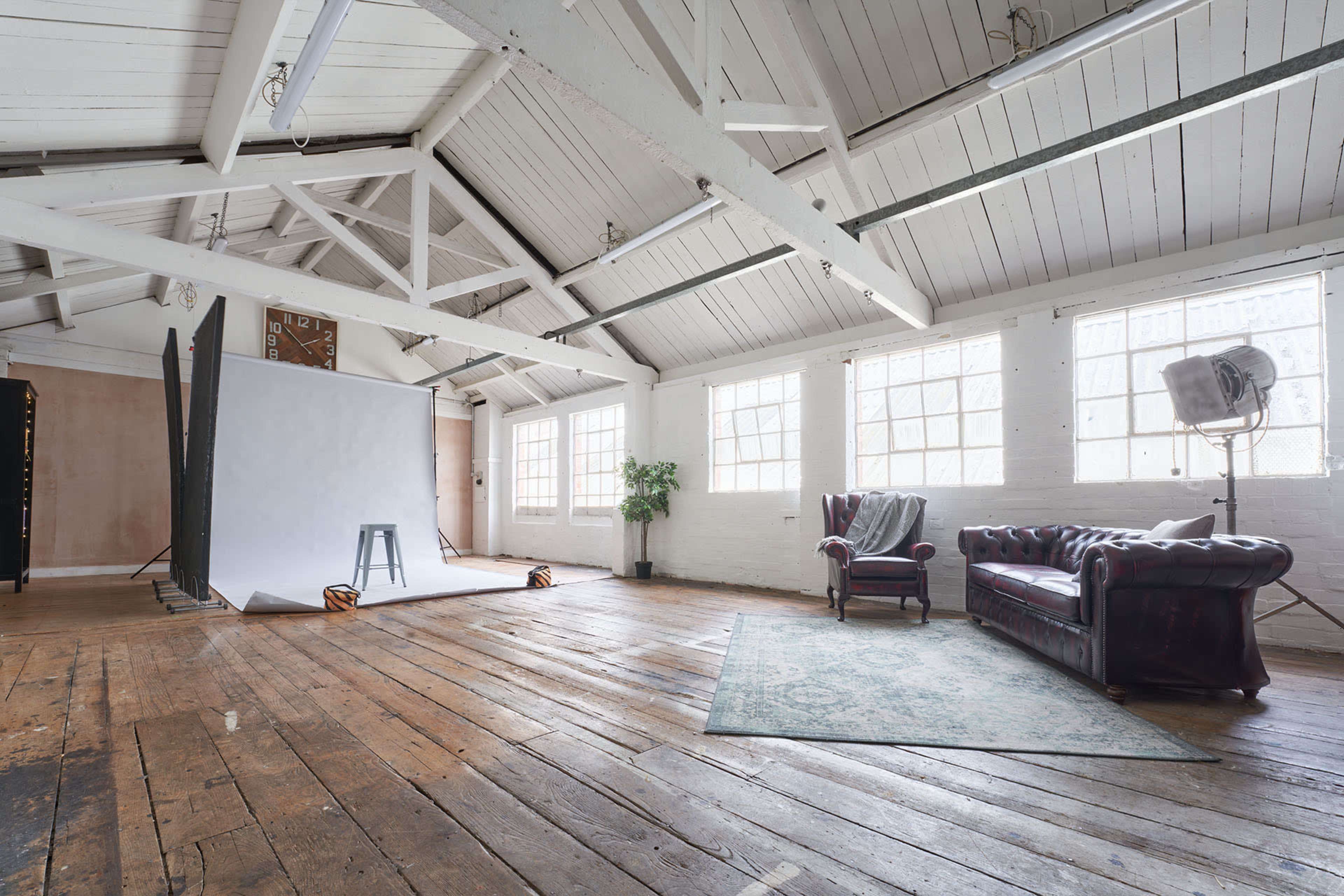 A spacious, well-lit studio with wooden floors, a white background setup, a dark leather couch, a stool, and a potted plant.