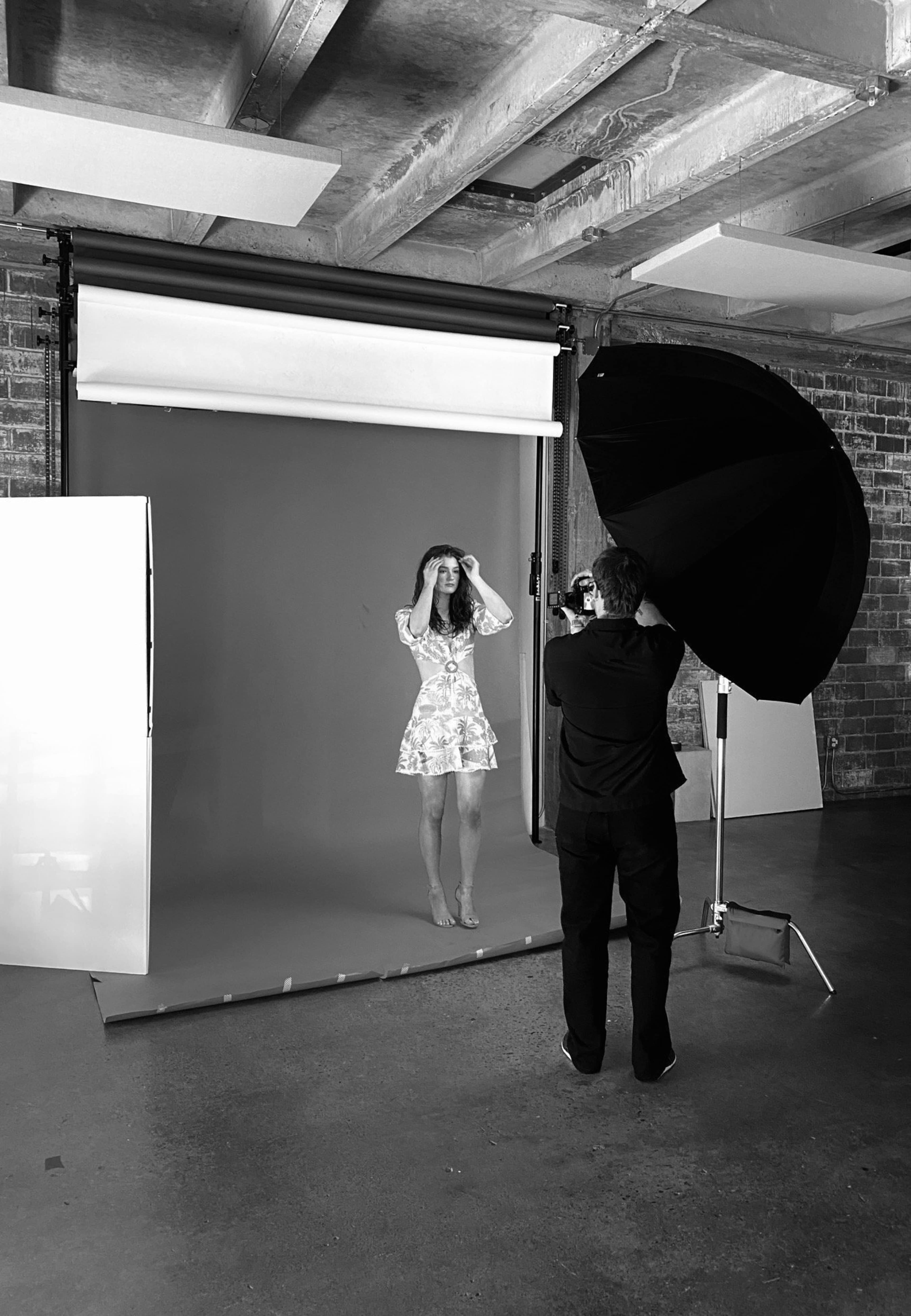A photographer is taking pictures of a model posing in a studio with a gray backdrop and an umbrella light.