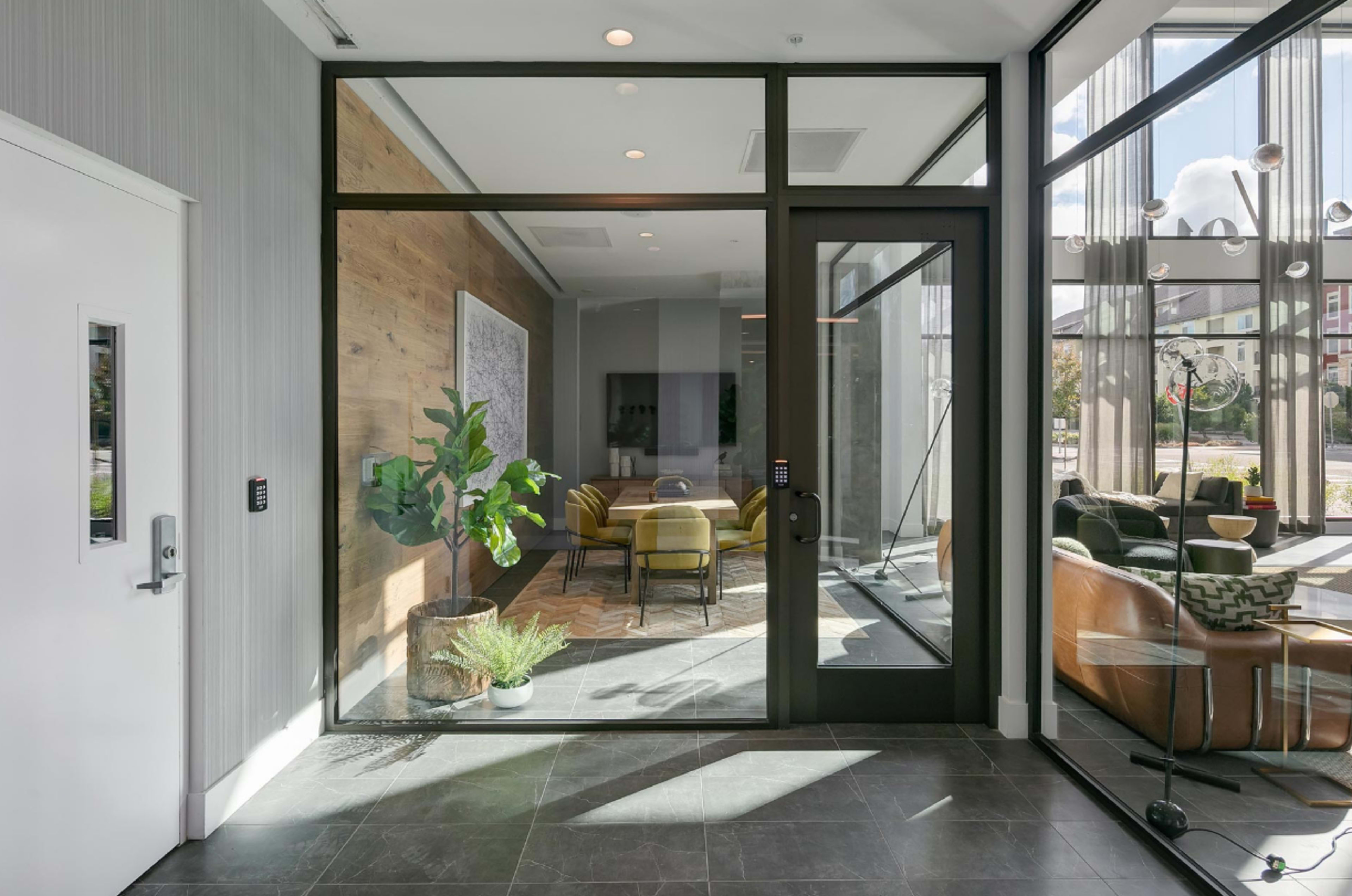 A modern entrance area with glass doors leading into a lounge featuring a wooden accent wall, seating, and decorative plants.