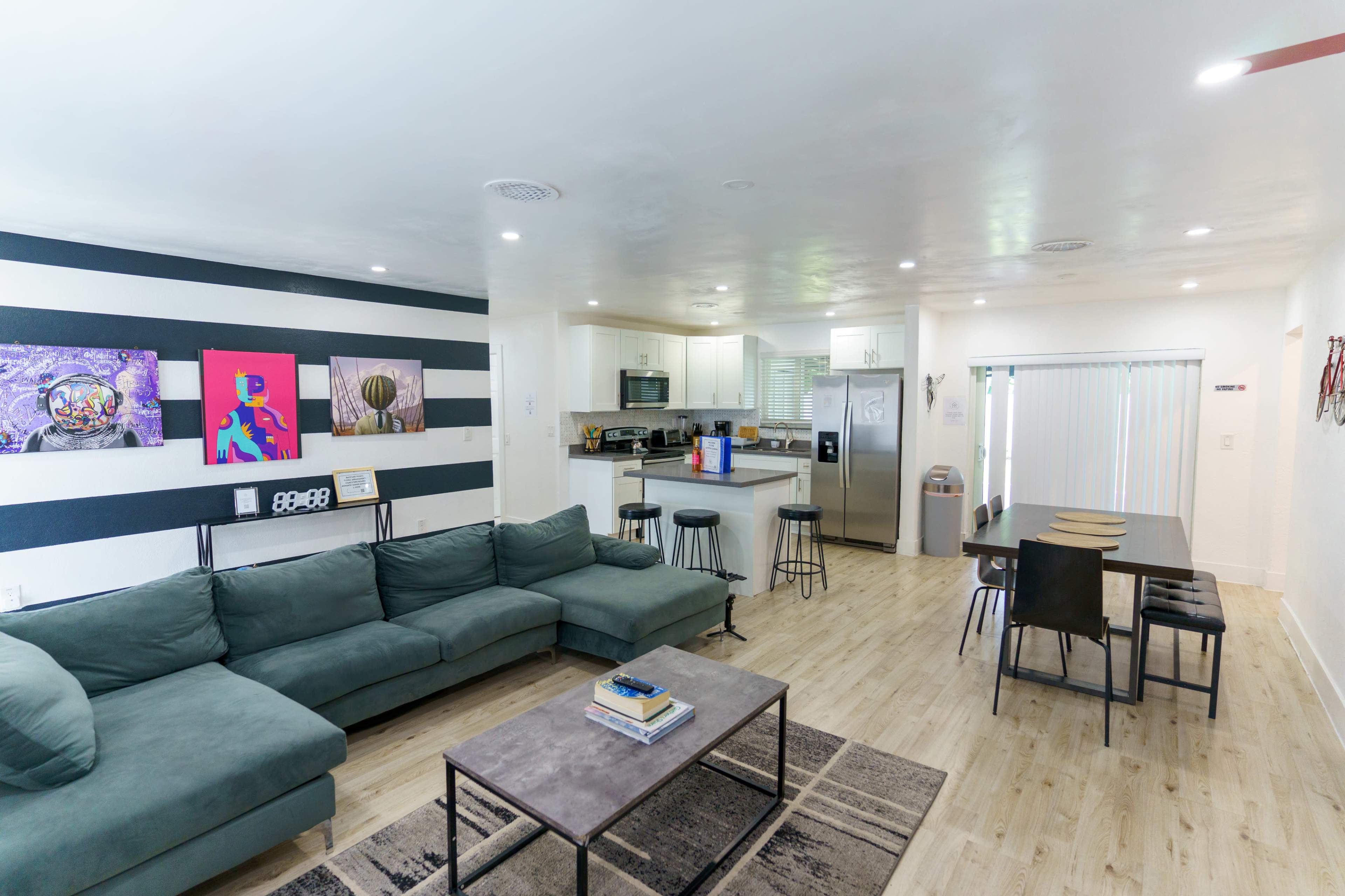 A modern open-concept living area featuring a green sectional sofa, a dining table, and a kitchen area with white cabinetry and stainless steel appliances.