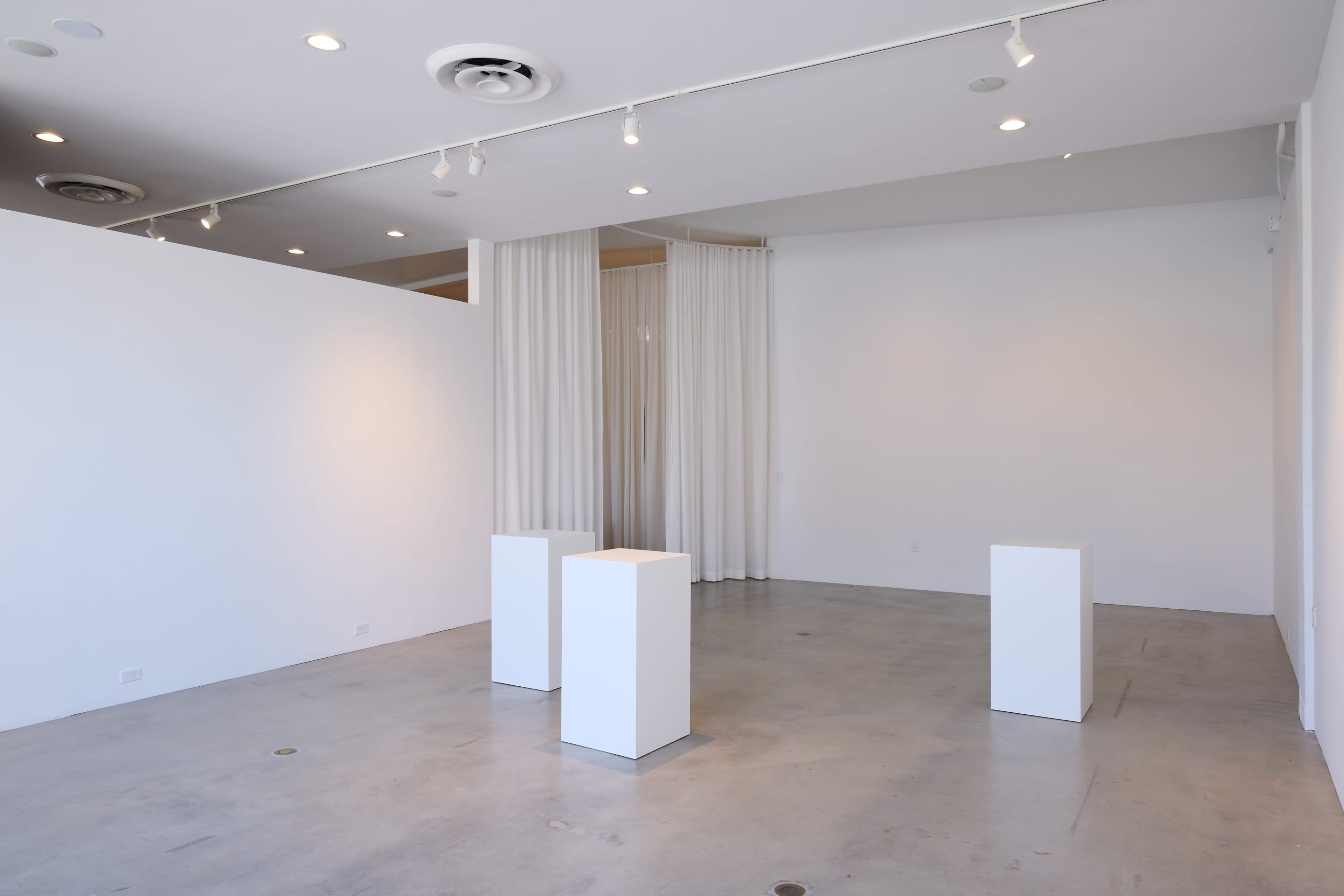 The image shows a minimalist gallery space featuring three white display pedestals on a concrete floor, surrounded by plain white walls and sheer curtains.