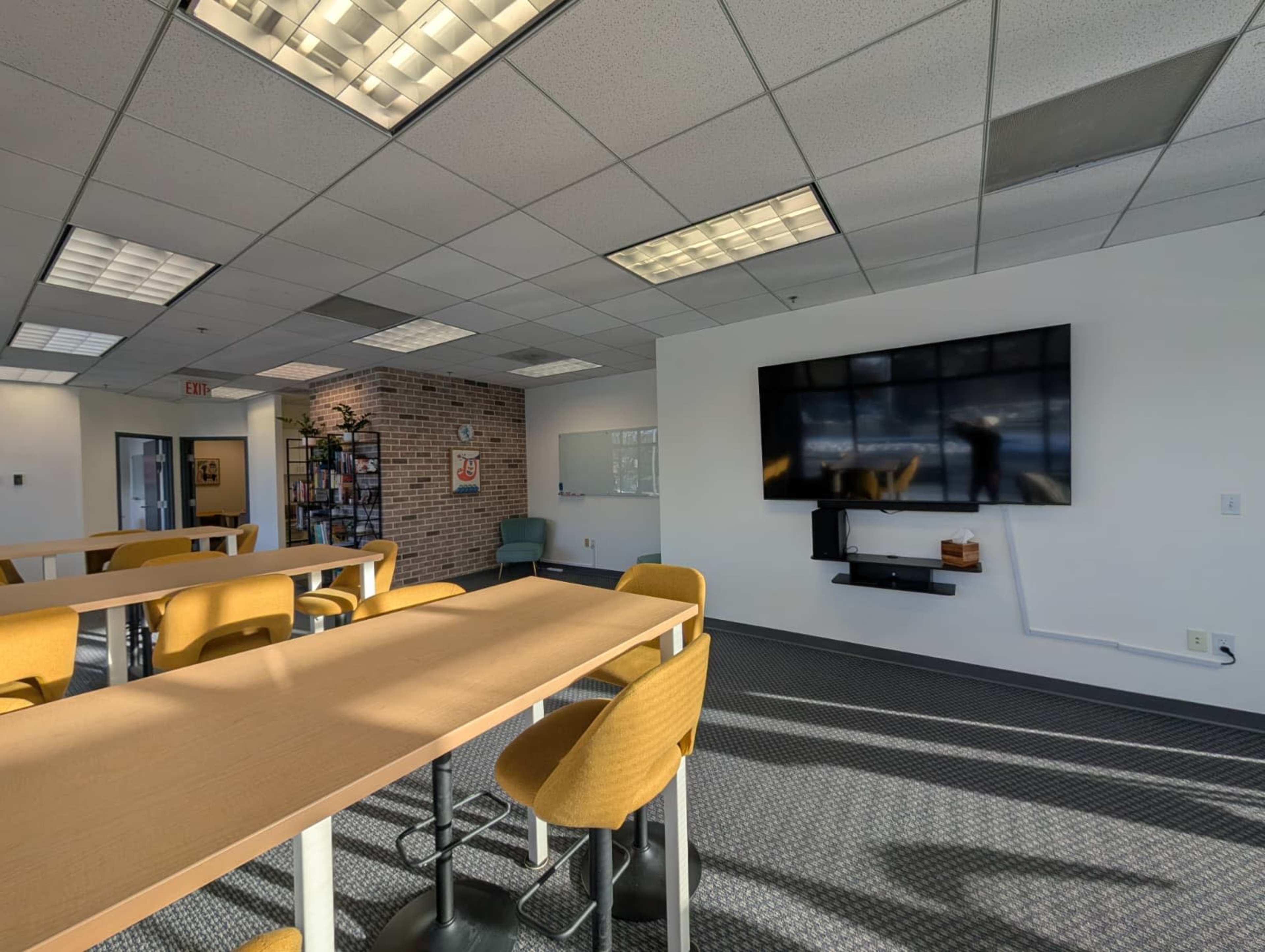 The image shows a bright office space featuring yellow chairs at a long table, a large wall-mounted TV, and a whiteboard on the wall.