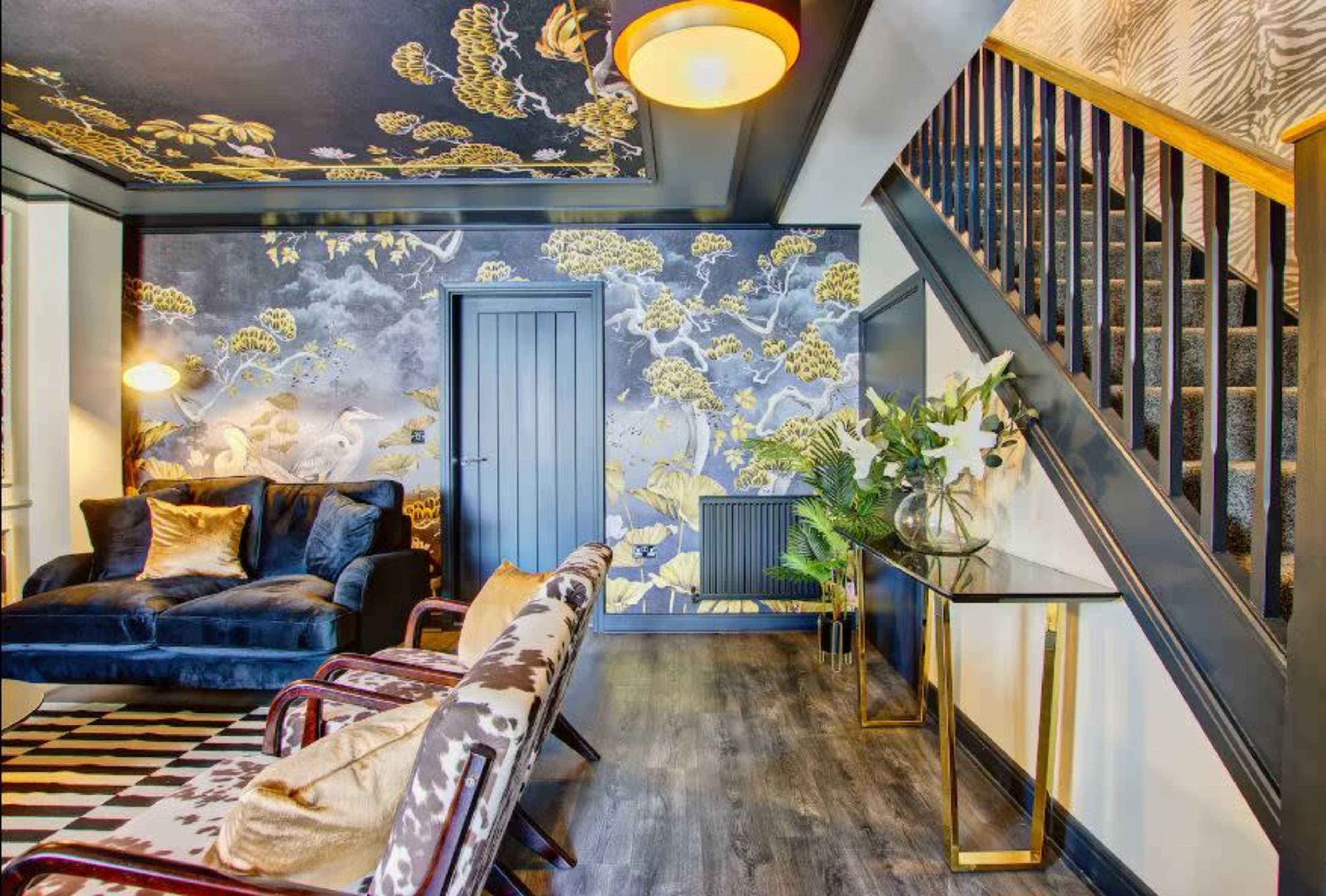 The image shows a stylish living room featuring a dark floral wallpaper, a navy blue couch, two patterned armchairs, and a staircase leading to an upper level.