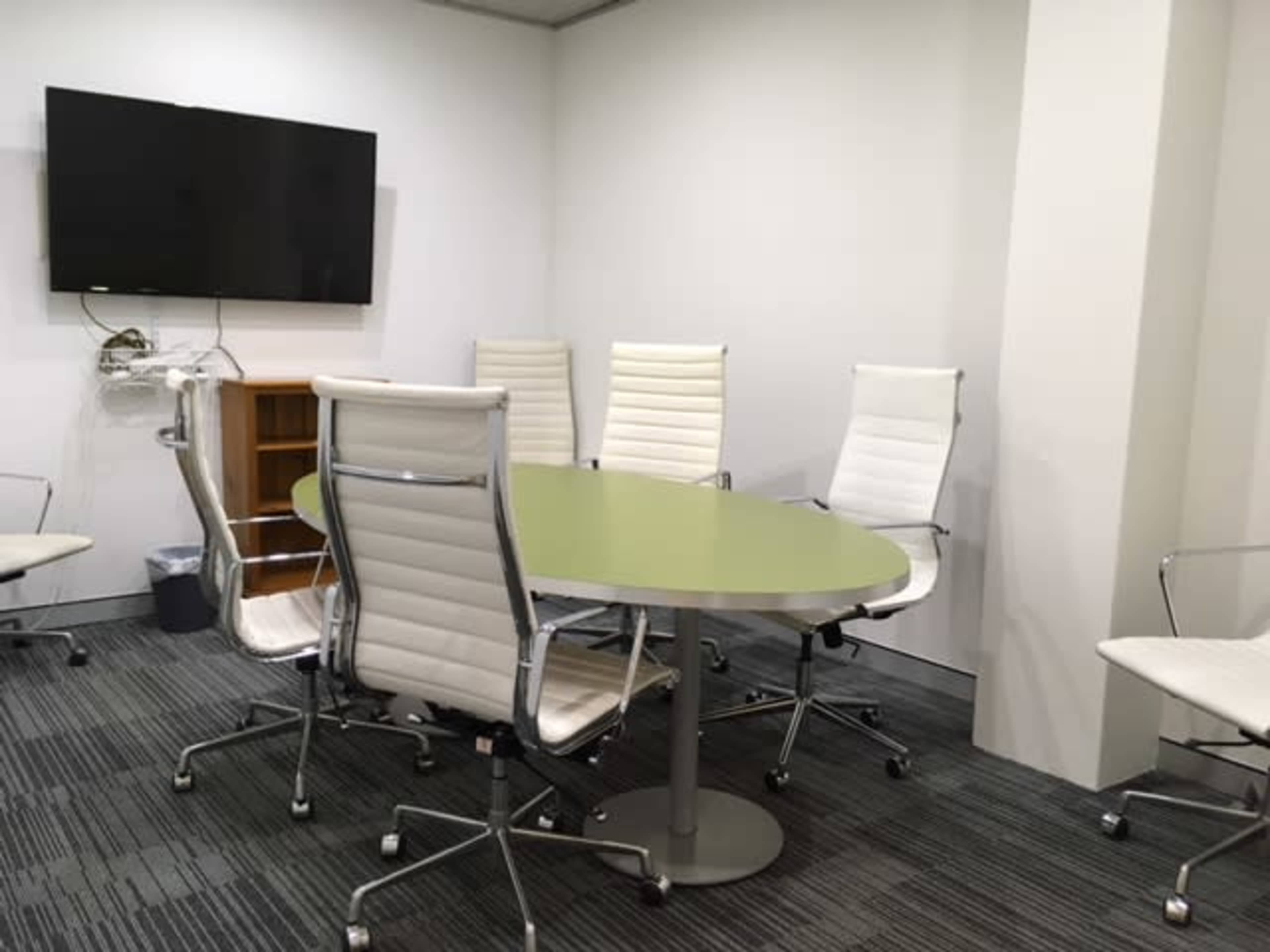A conference room features a green oval table surrounded by six white ergonomic chairs and a wall-mounted television.