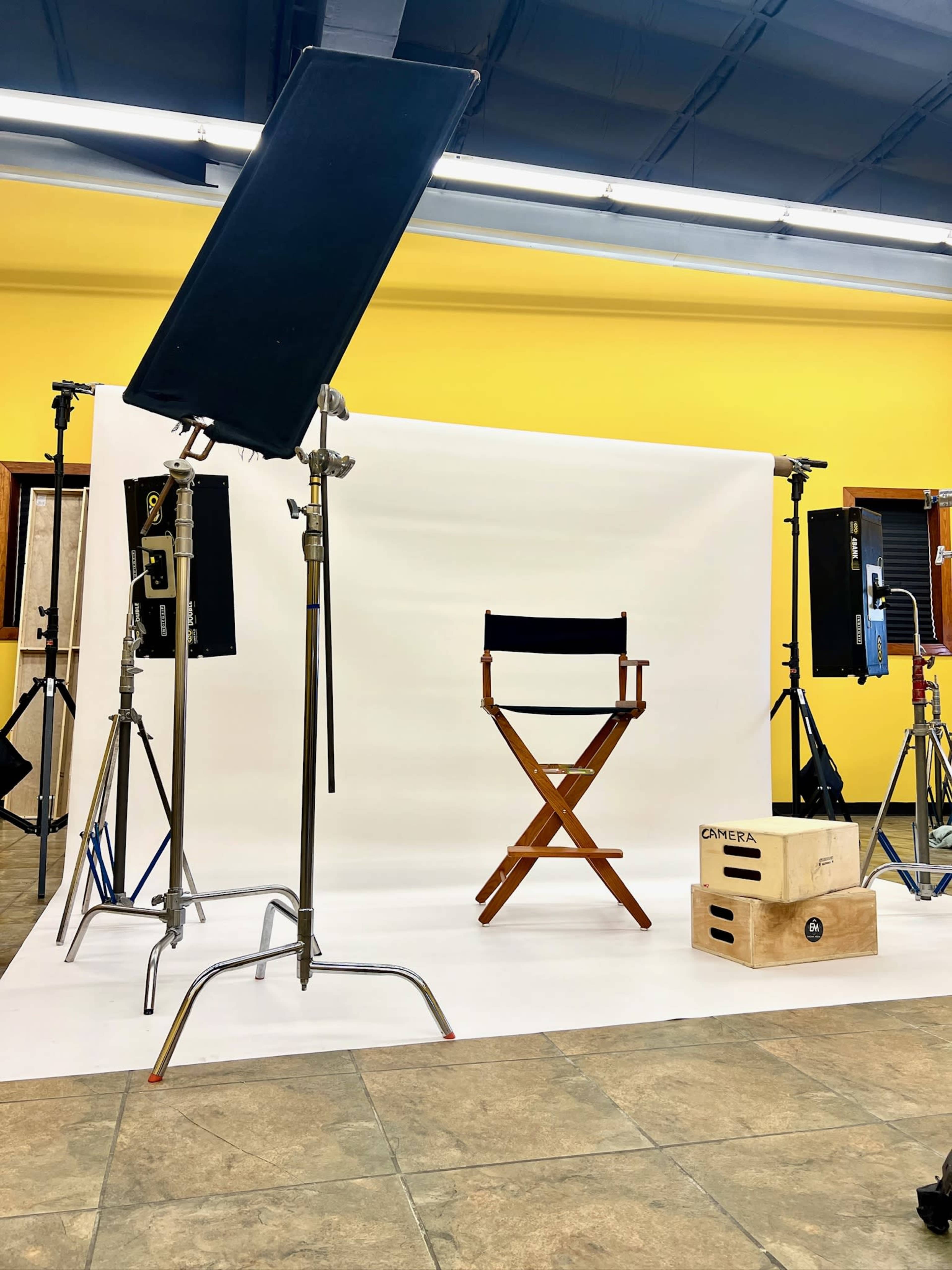The image shows a photography studio setup with a director's chair, lighting equipment, and a plain white backdrop.