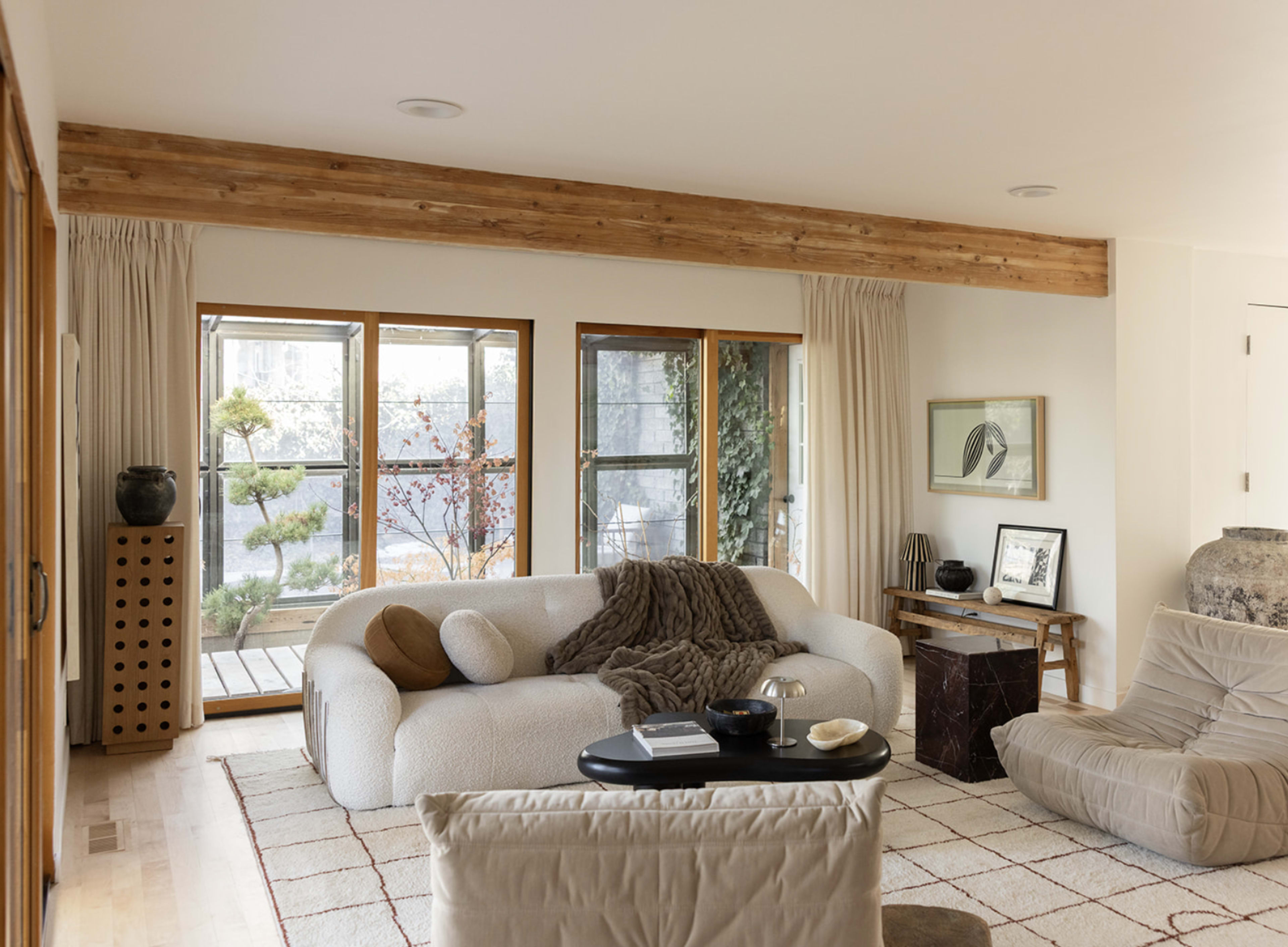 The image depicts a modern living room with a curved white sofa, a small black table, and large windows overlooking a garden.