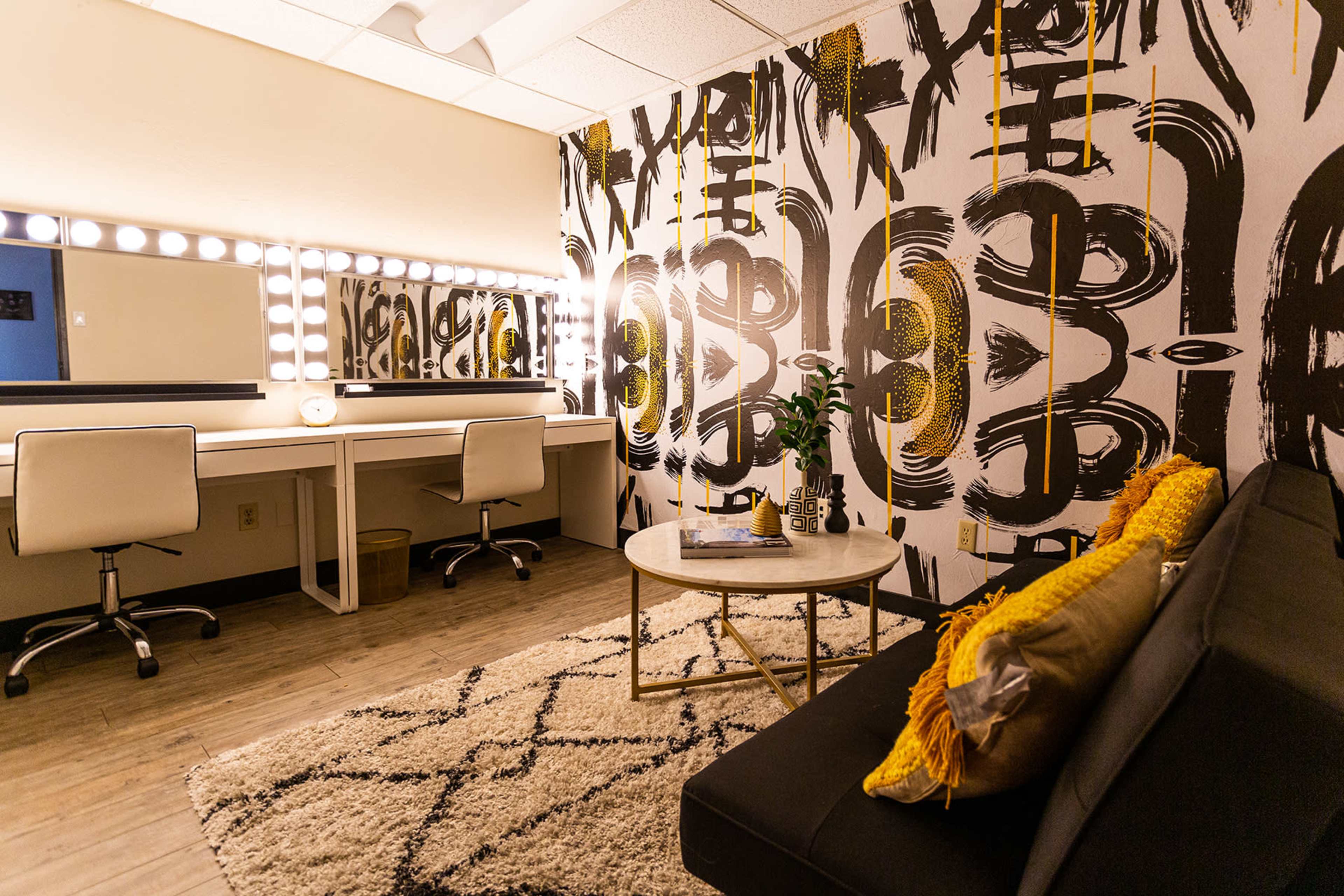 A stylish dressing room with a large illuminated mirror, a gray couch, a circular coffee table, and a patterned wall design.